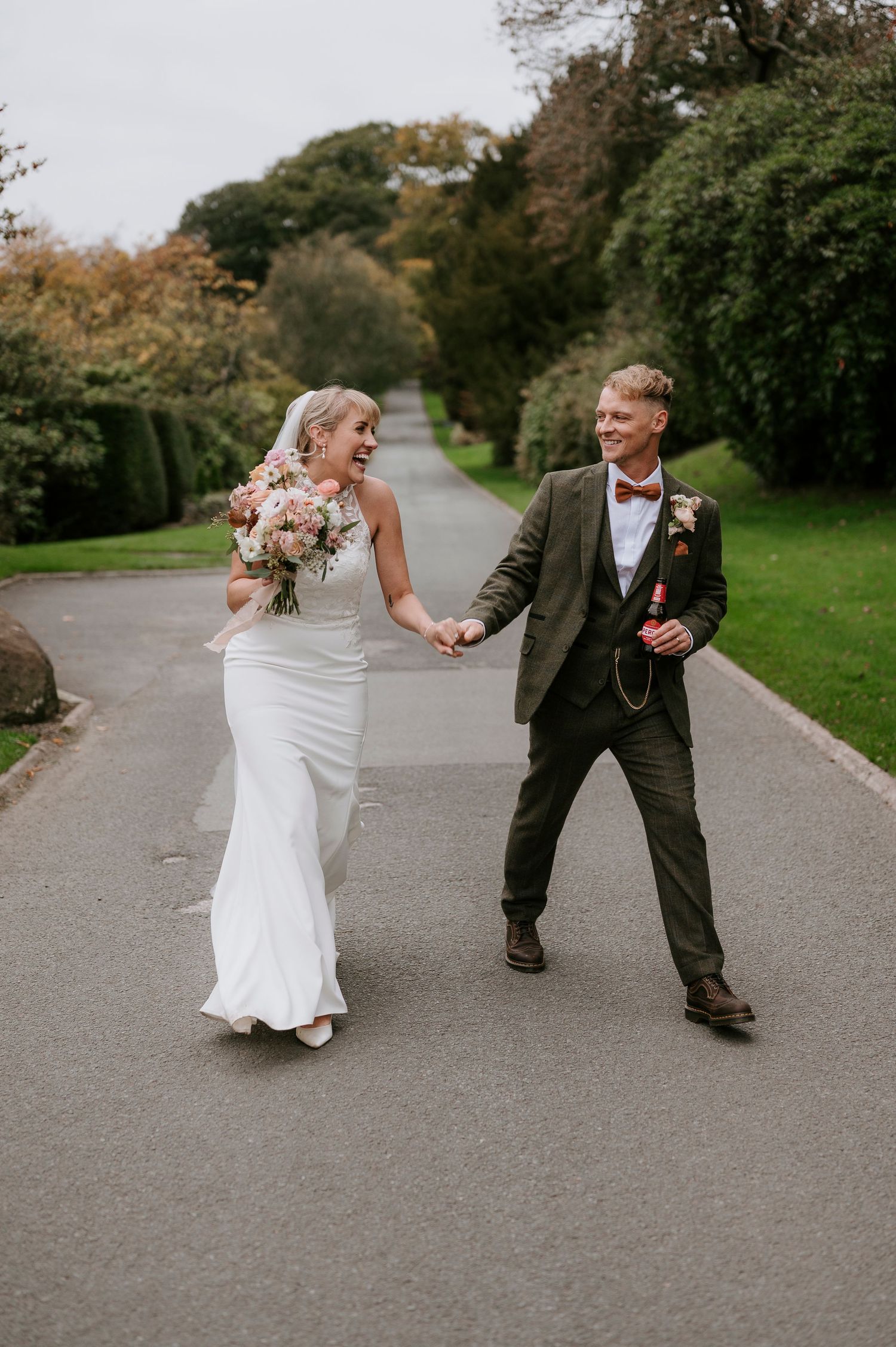 Couple in wedding attire walking and laughing together on a paved path surrounded by greenery.