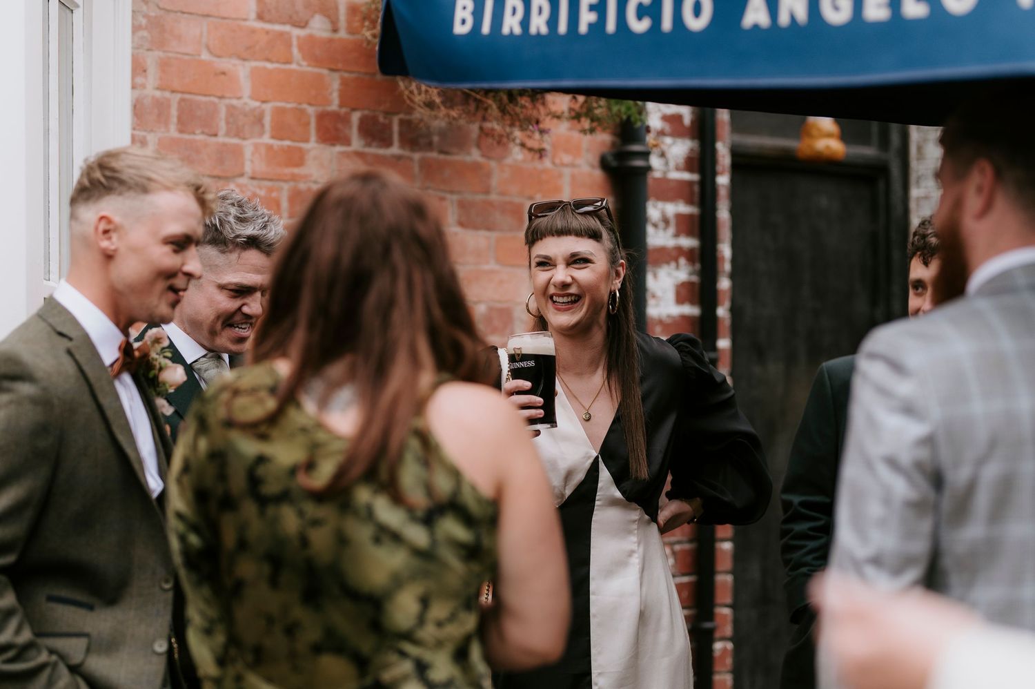 Wedding guests gather outside a brick building with Birrificio Angelo signage for an outdoor celebration.