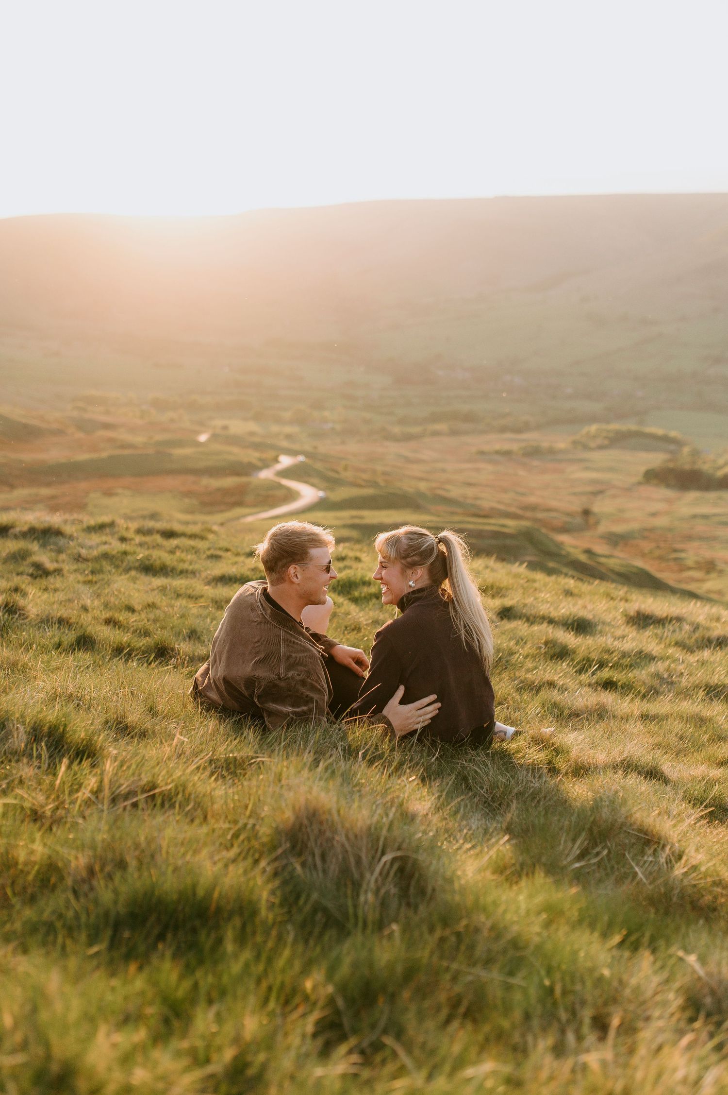A couple lies together on a grassy hillside overlooking a winding road through misty countryside at sunset.