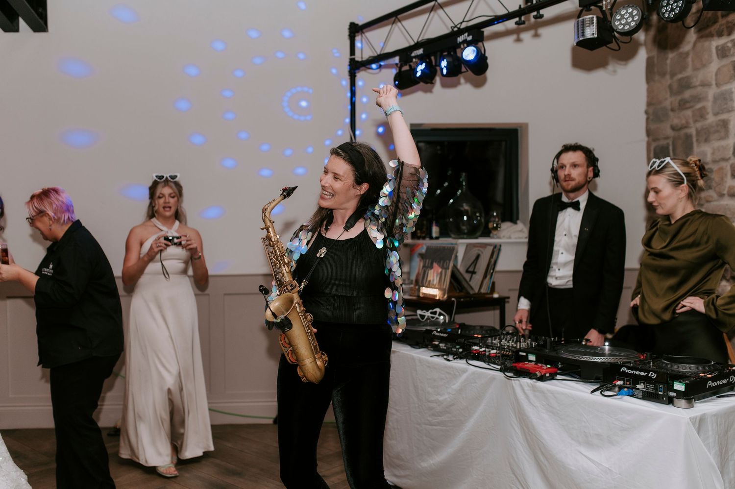 A saxophone player performs at a wedding reception with blue lighting and DJ equipment visible in the background.
