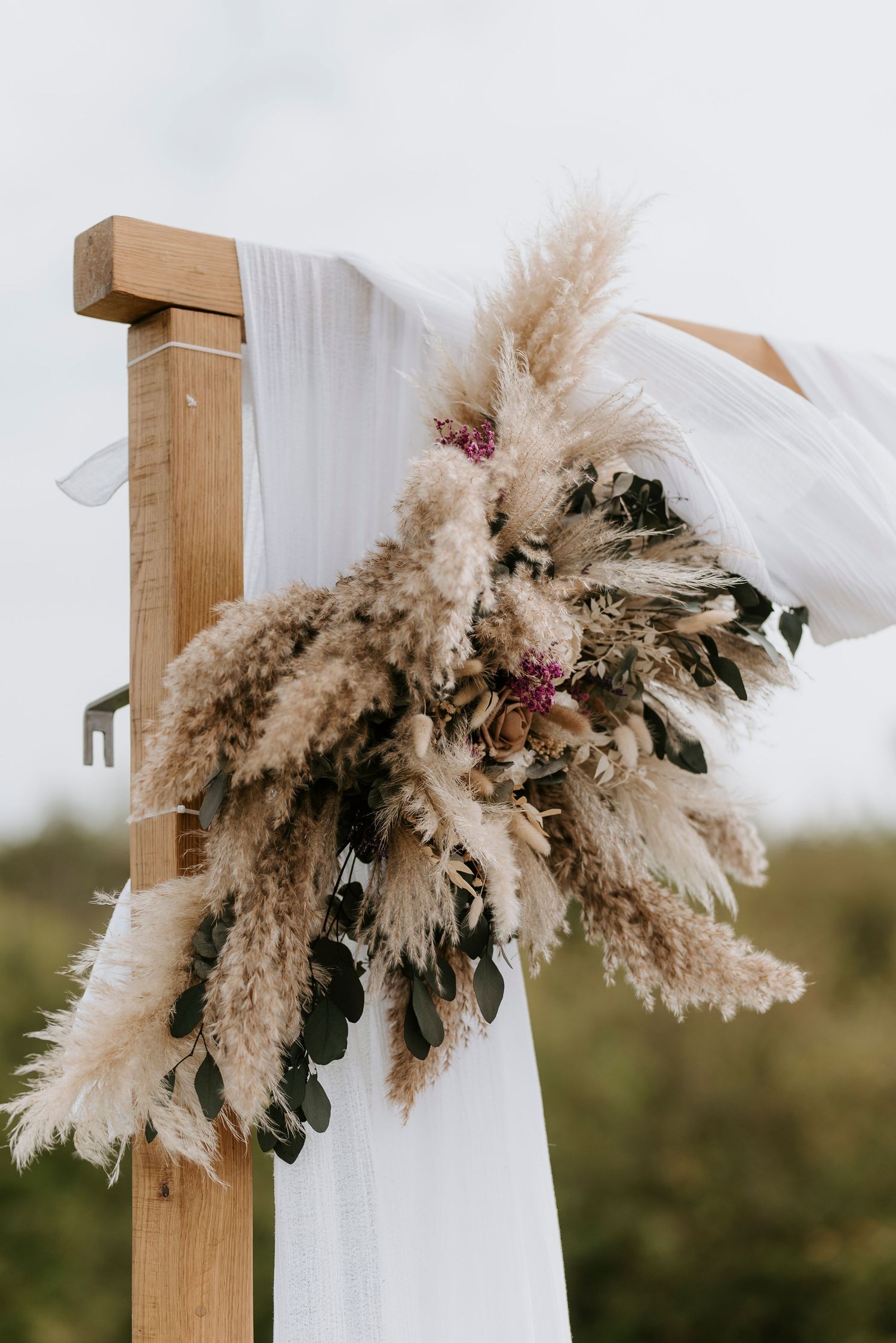 Boho wedding arch decorated with flowing white fabric, pampas grass, and dried floral arrangements in a natural outdoor setting.