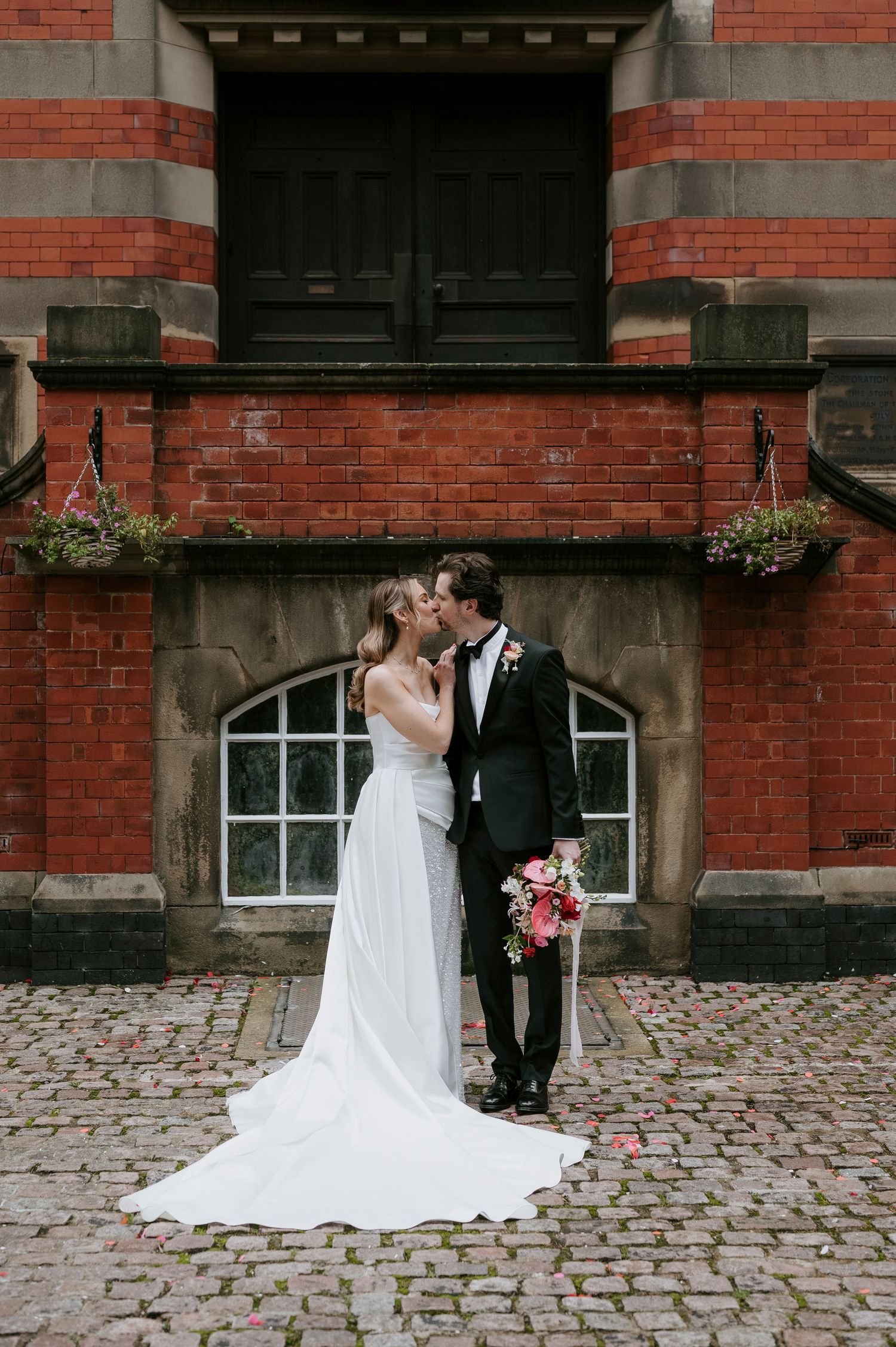 A romantic wedding couple shares a kiss against a red brick building with cobblestone pavement.