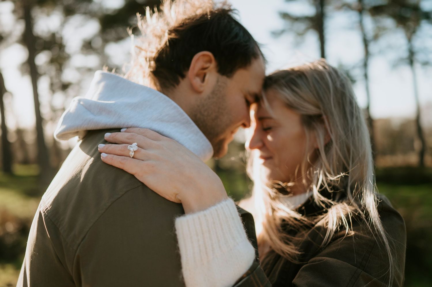 Romantic couple embraces outdoors in warm sunlight with trees and nature in background.