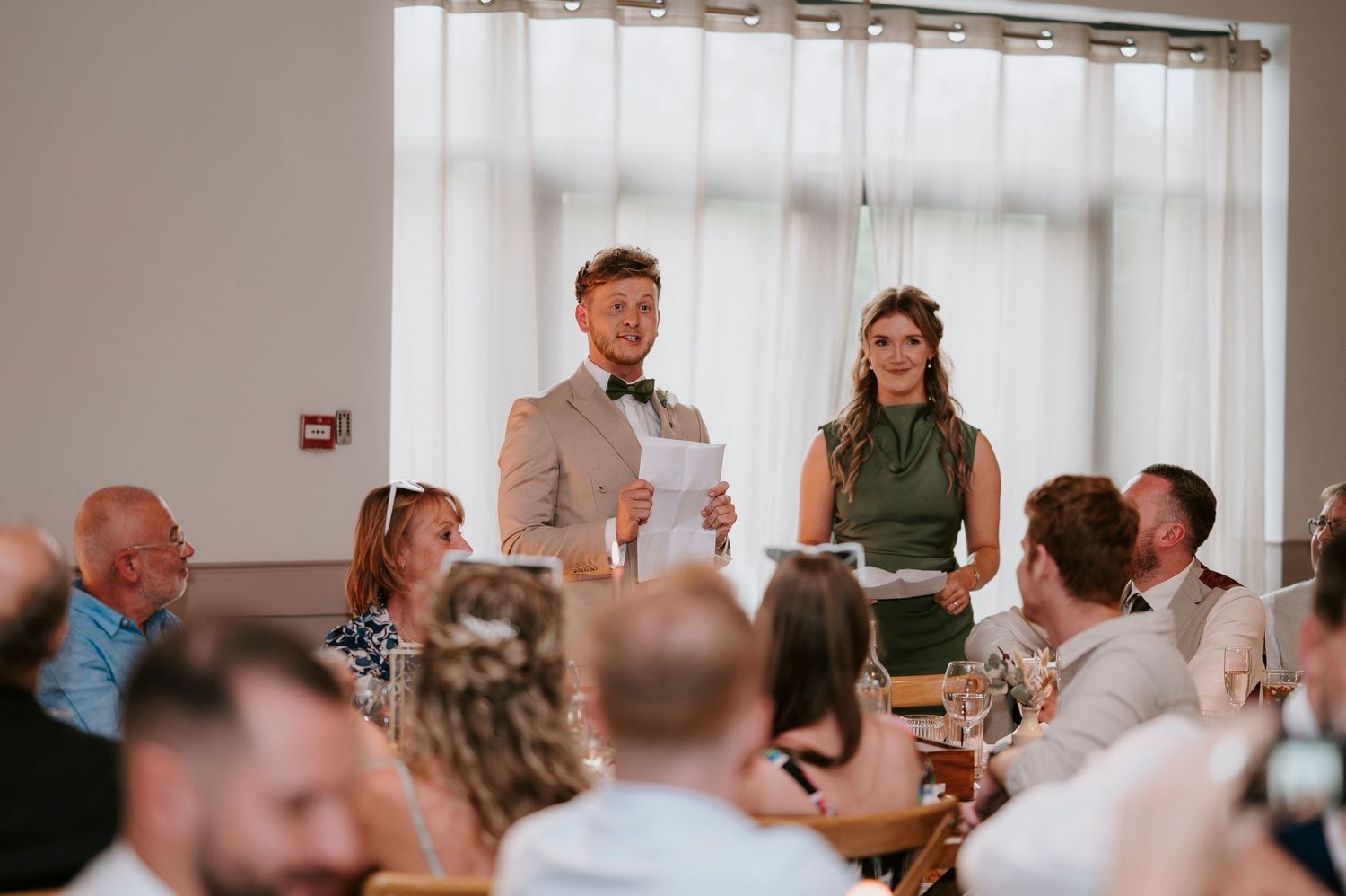 Wedding guests seated at long tables in a bright reception hall raise their glasses in a celebratory toast.