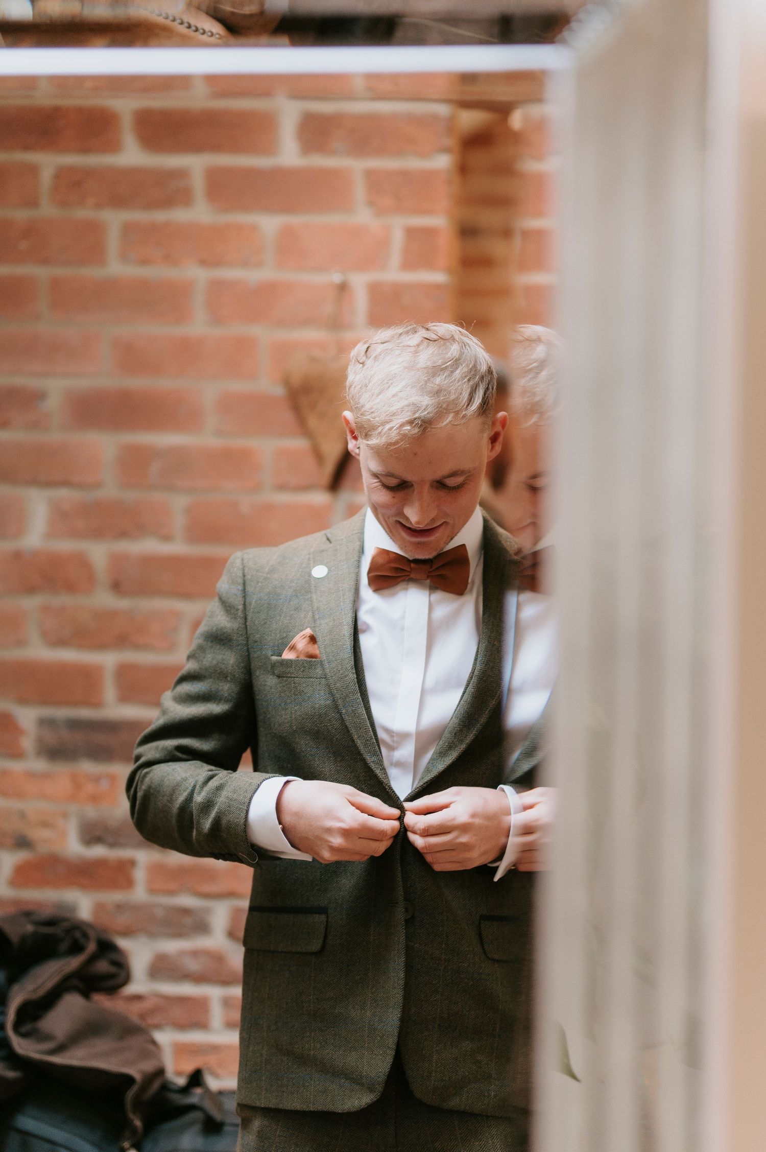 A person in a gray blazer and white shirt adjusts their collar while standing against a brick wall.