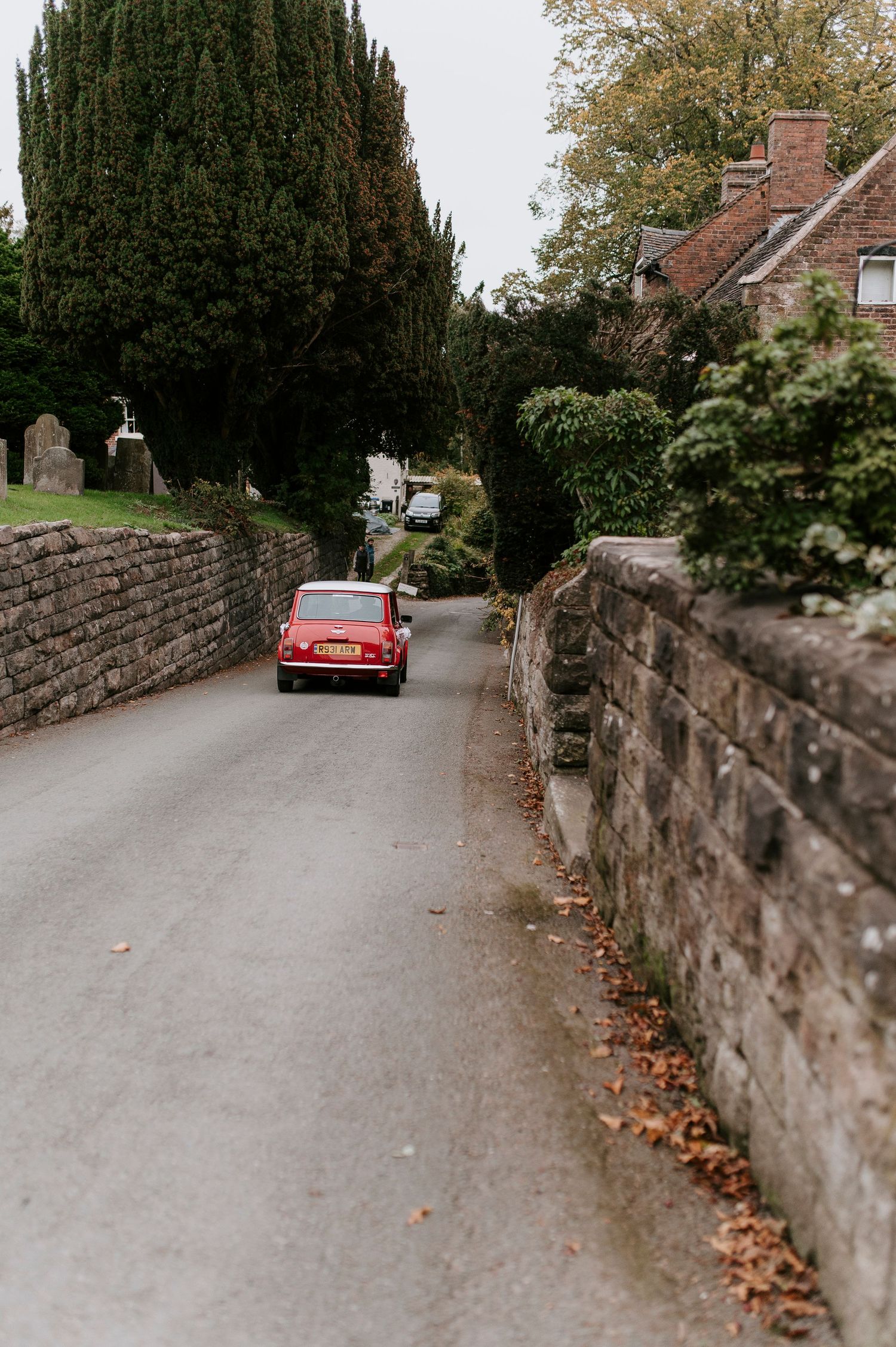 A red car drives along a narrow road bordered by an old stone wall with autumn leaves scattered on the ground.