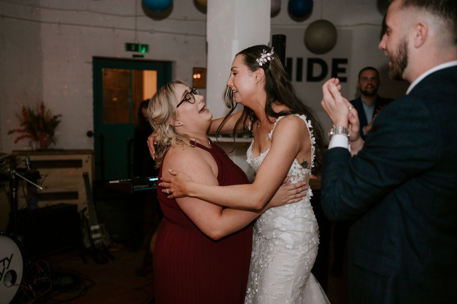 Bride in lace gown sharing emotional dance with guest in burgundy dress at wedding reception.
