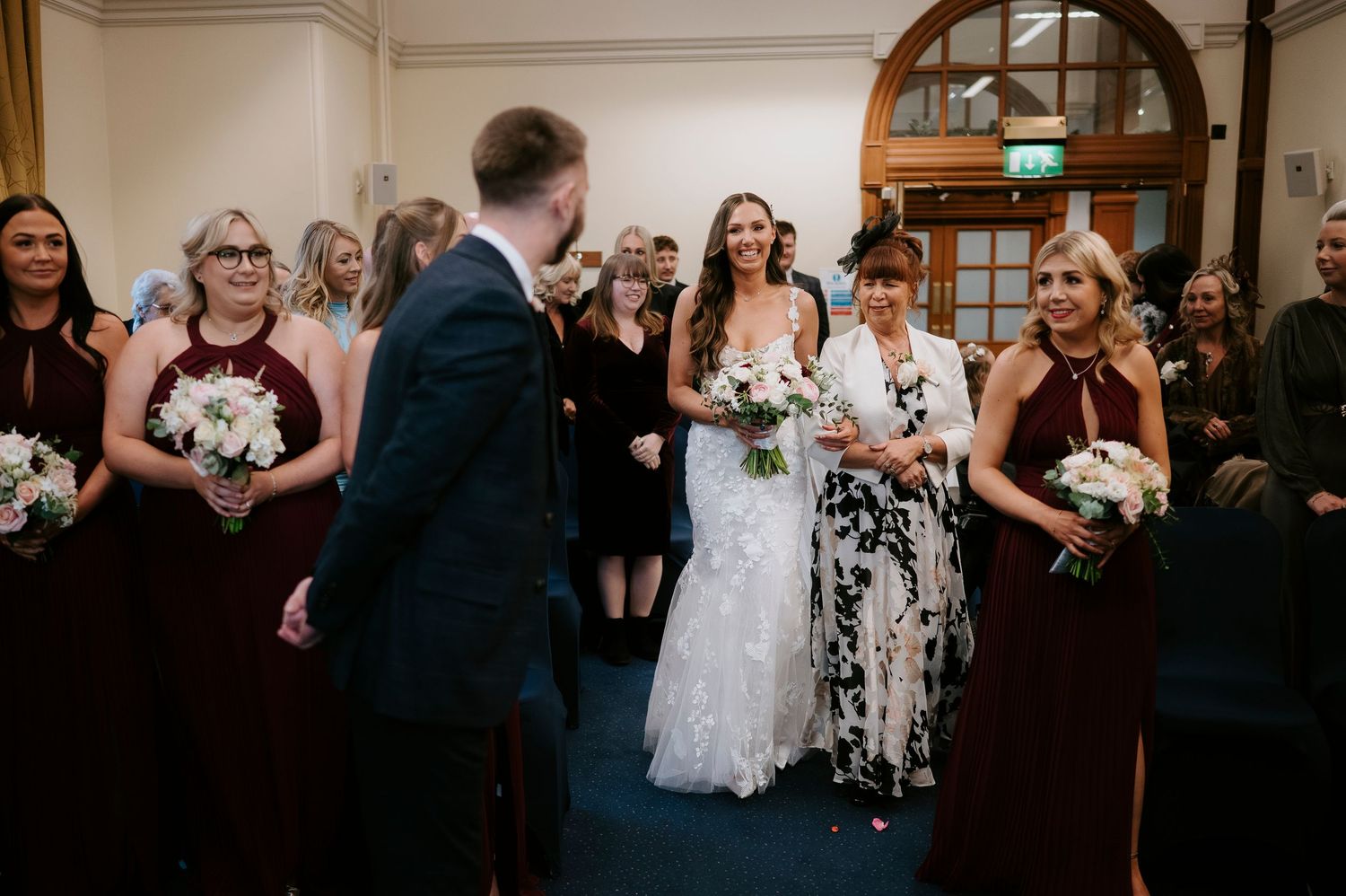 Bride walks down the aisle in lace gown with bridesmaids in burgundy dresses at elegant indoor wedding ceremony.