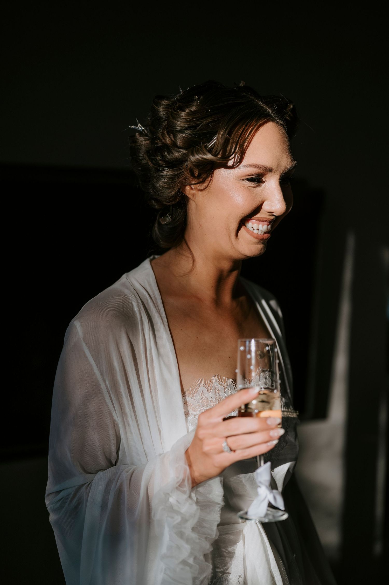 Smiling bride in white robe holding champagne glass during wedding morning preparations.