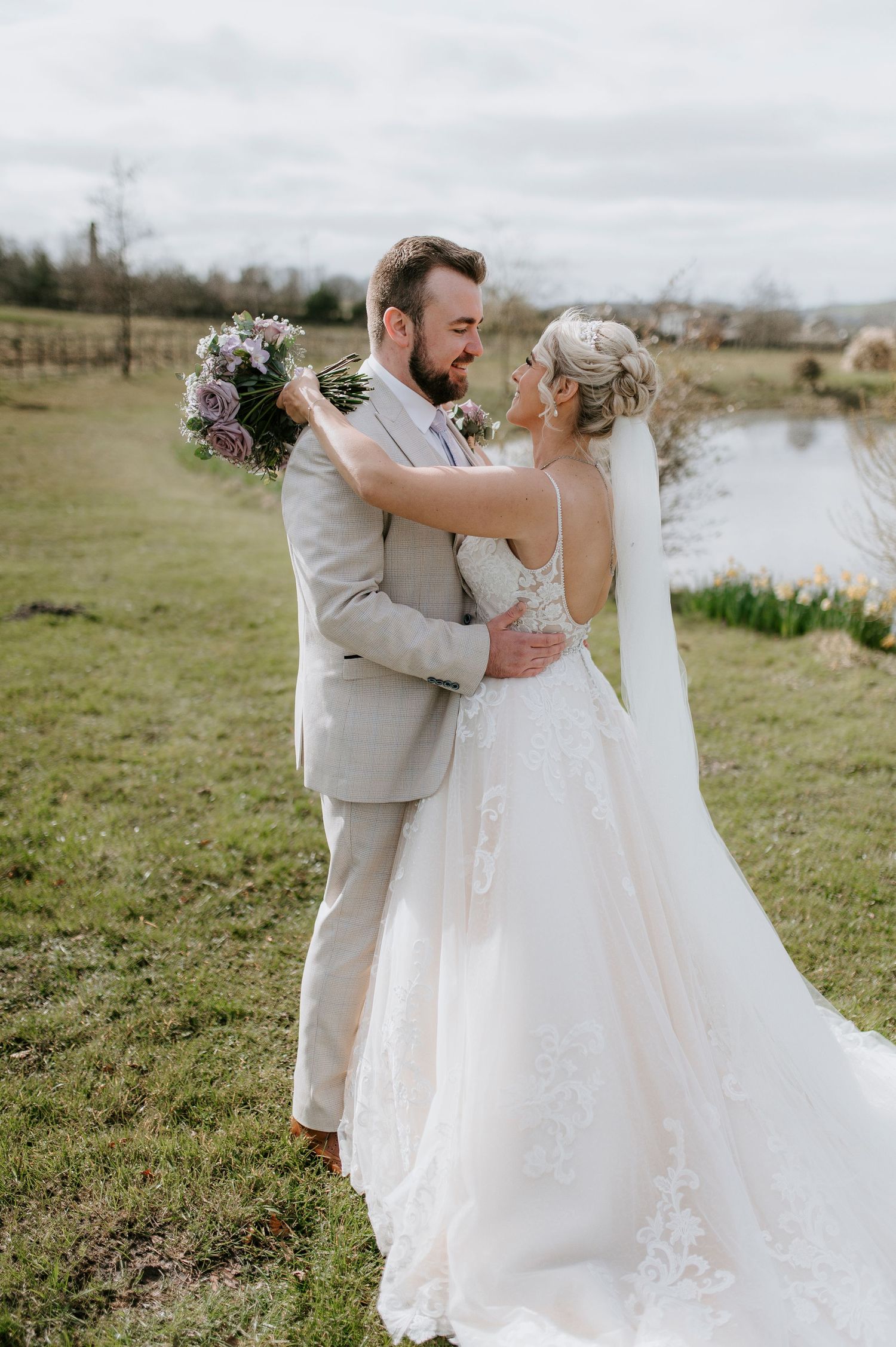 A couple embraces outdoors at their wedding, with the bride in a lace gown and groom in light gray suit near a pond.