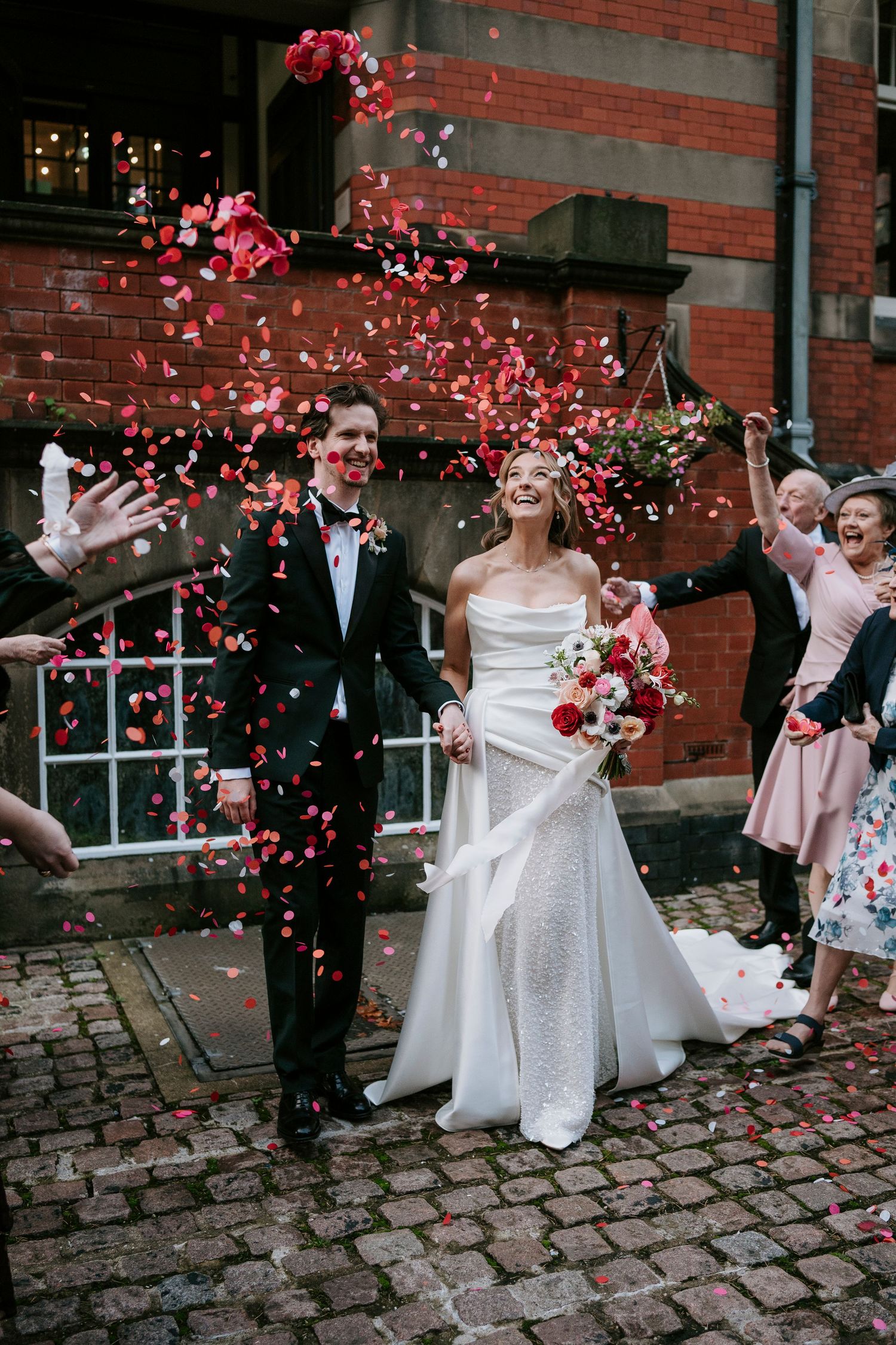 Wedding party celebrates with red flower petals being tossed in front of historic brick building.