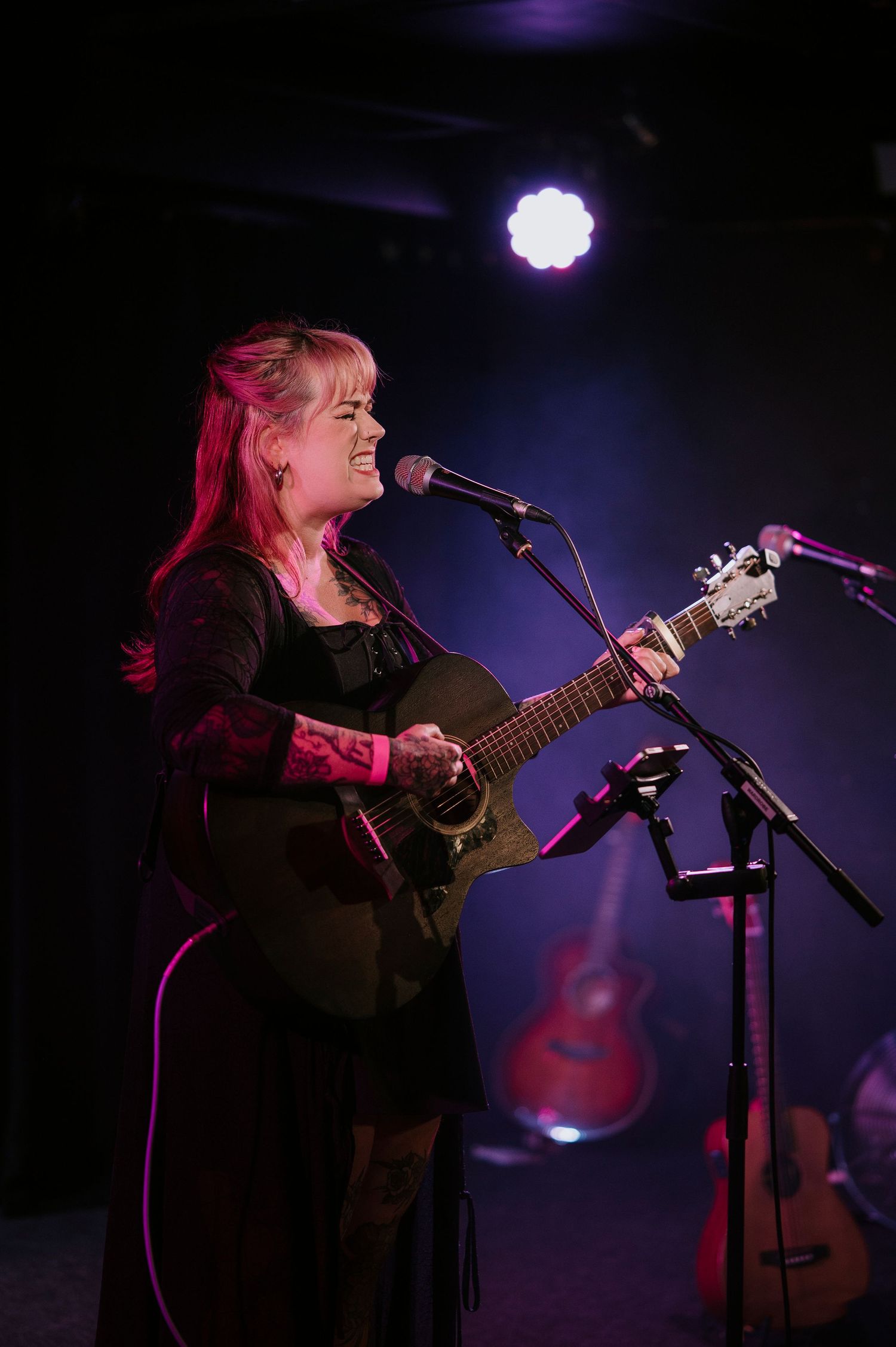 A musician plays acoustic guitar on a dark stage illuminated by purple spotlights and stage effects.