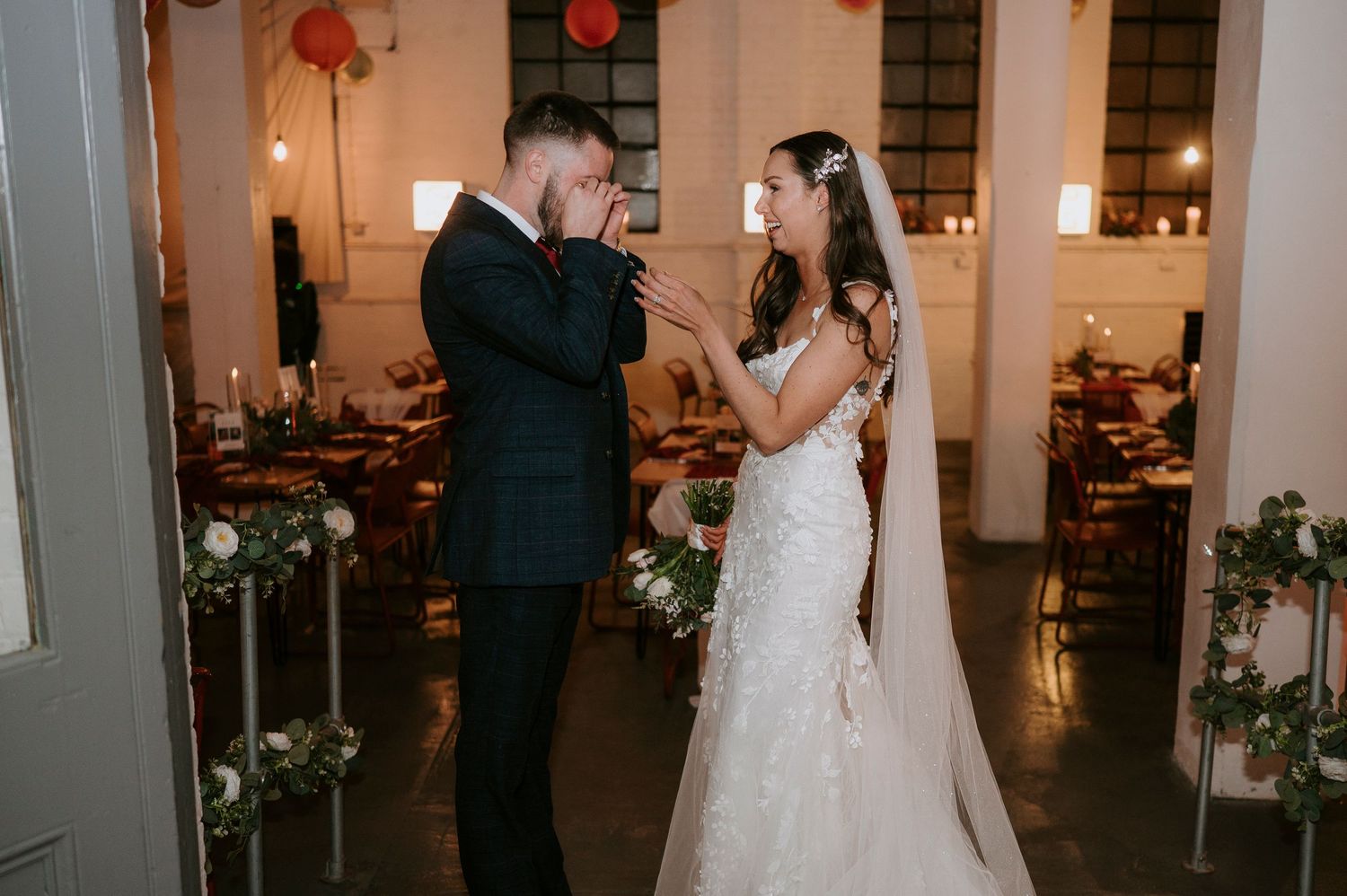 Emotional groom kisses bride's hand during first look moment in decorated industrial wedding venue with red accents.