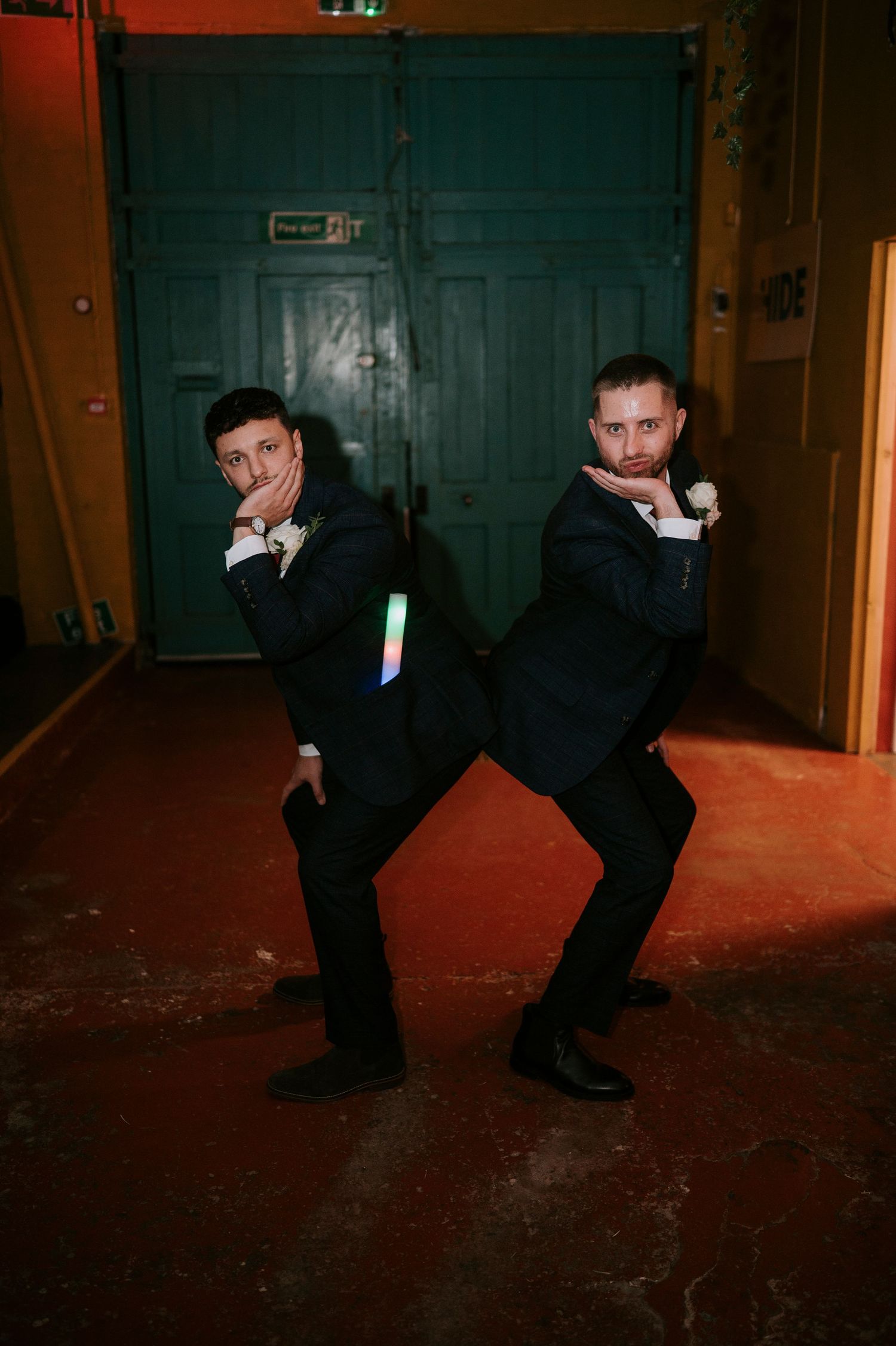 Two groomsmen in navy suits striking fun back-to-back pose with glow sticks at wedding reception.
