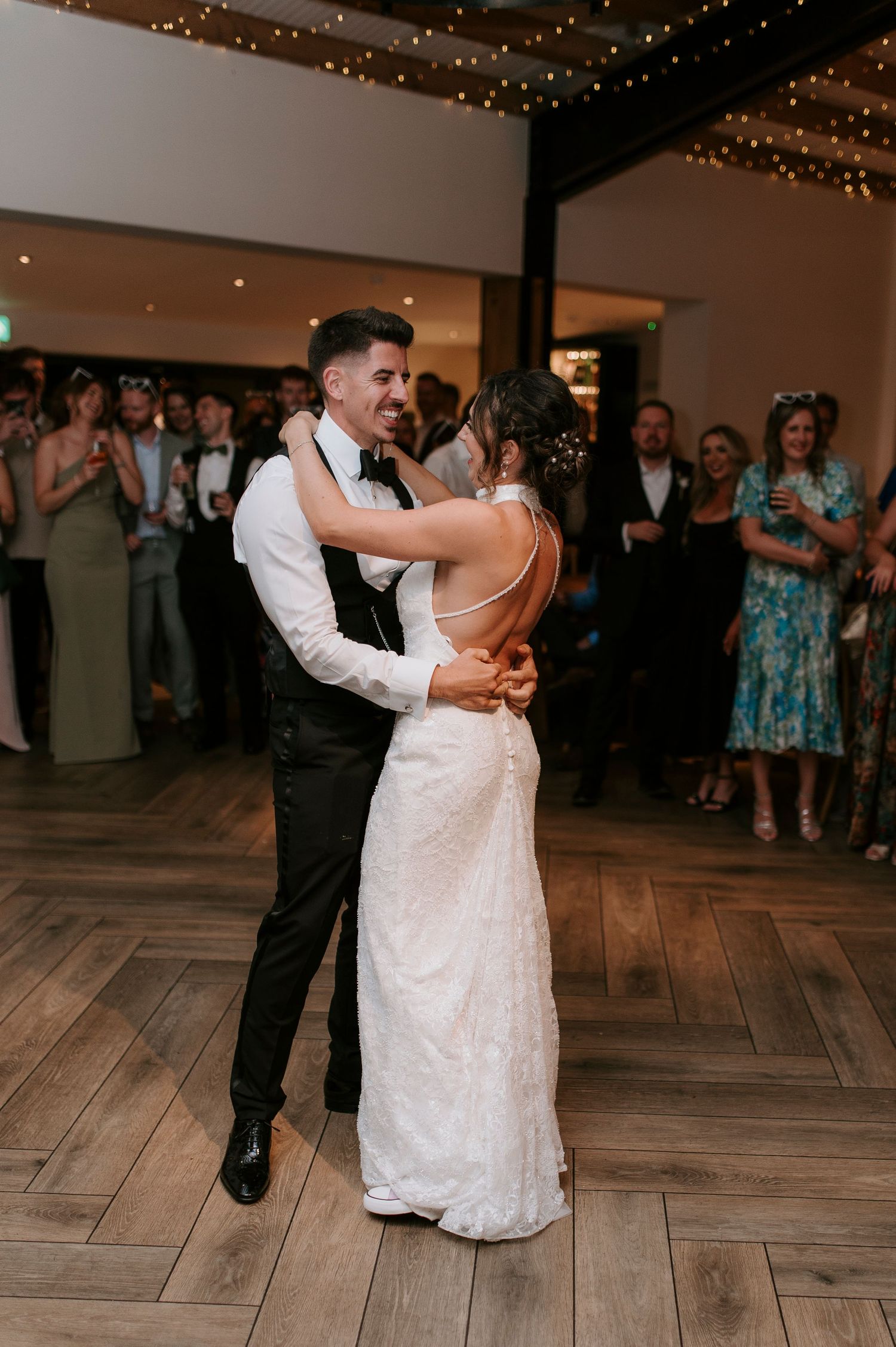A couple dances together on a wooden floor at a wedding reception with twinkle lights and guests in the background.