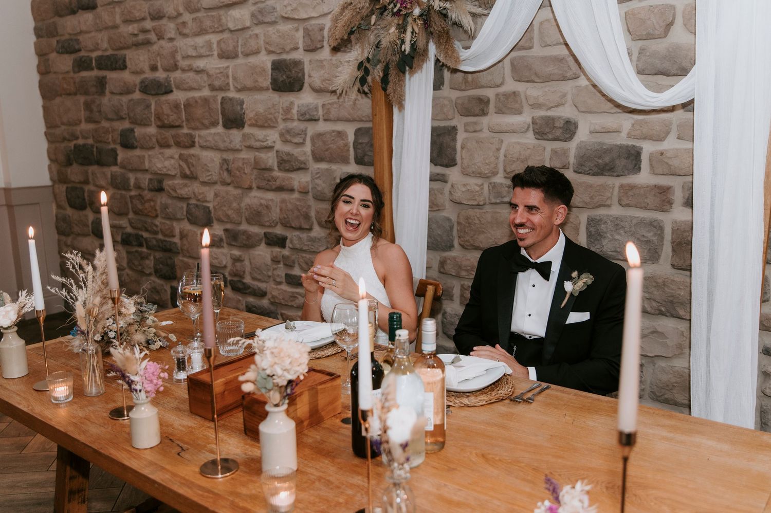 Wedding guests seated at wooden tables with brick walls and white decorative elements in a rustic reception venue.