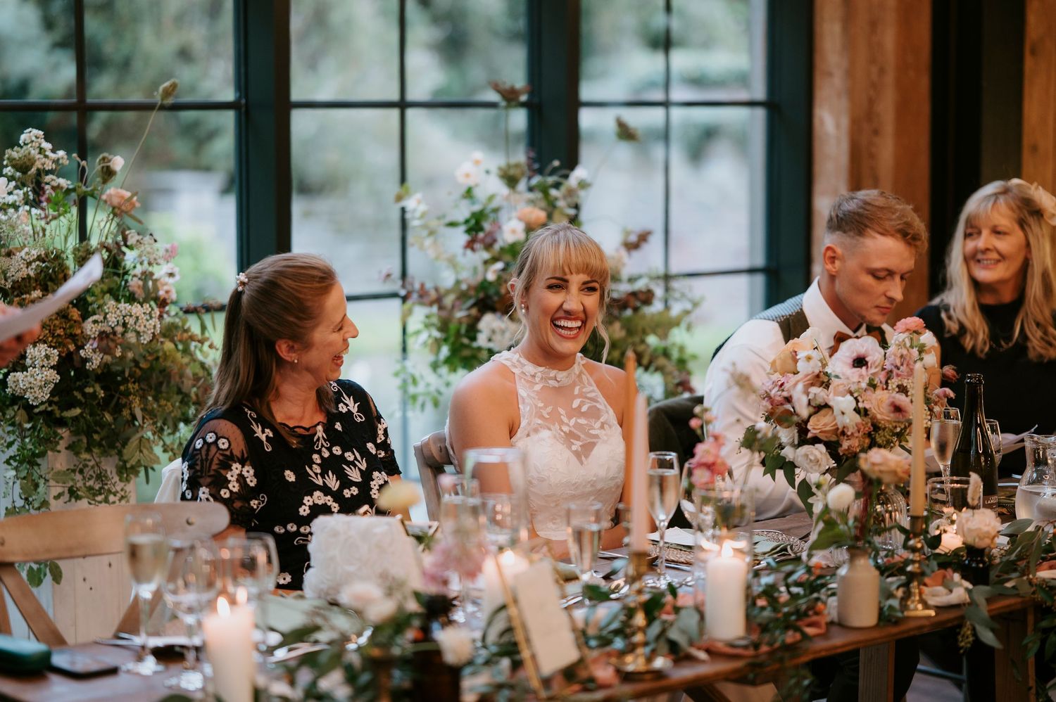 Wedding reception guests share joyful laughter around an elegantly decorated dining table with floral centerpieces.