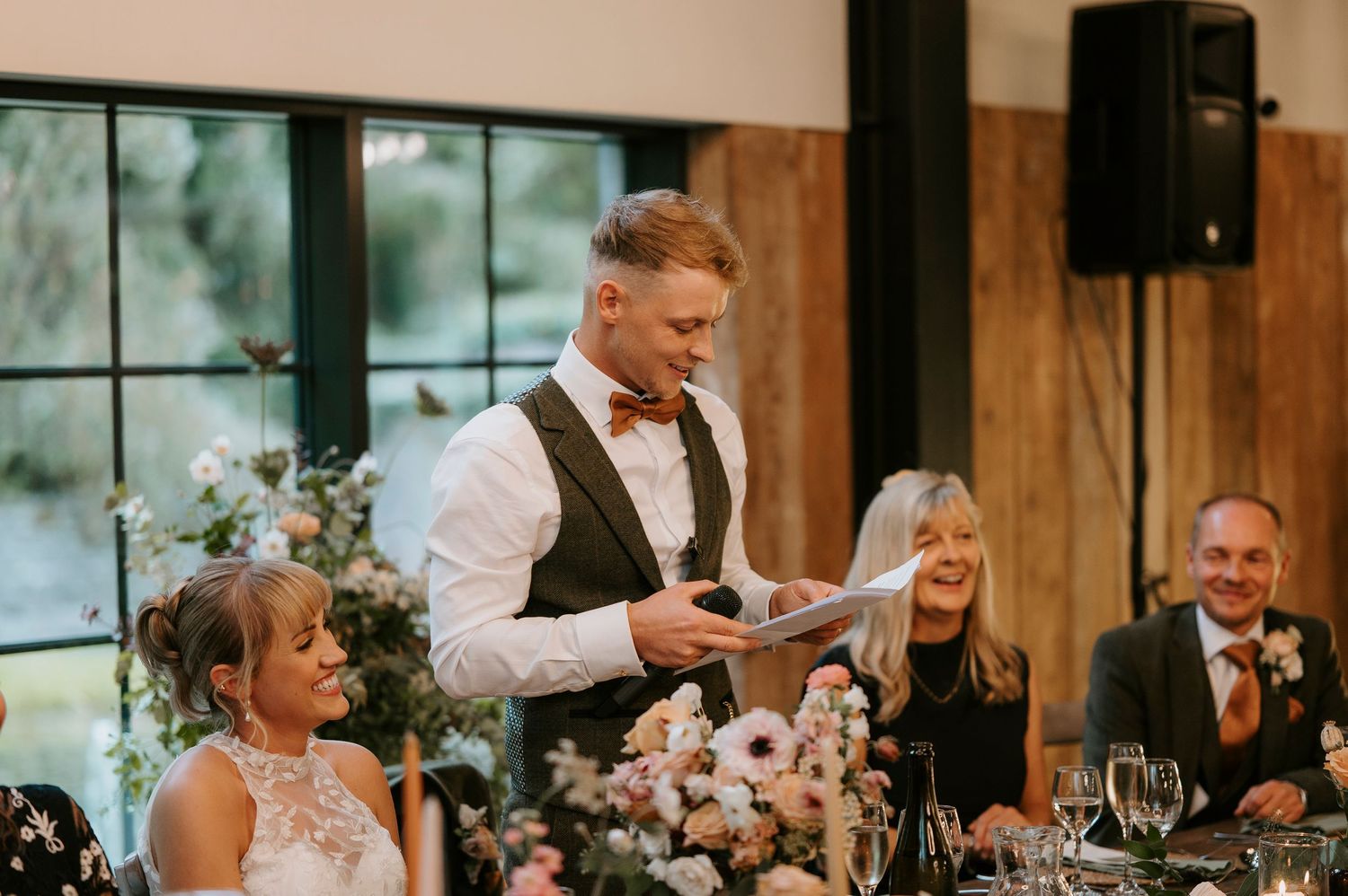 Wedding guests seated at reception tables share joyful reactions during speeches at an elegant indoor venue.