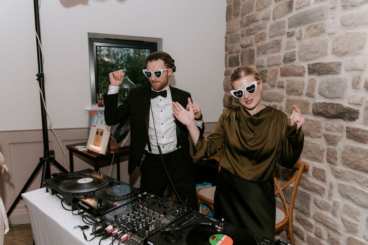 DJs performing at a party with DJ equipment and exposed brick walls in the background.