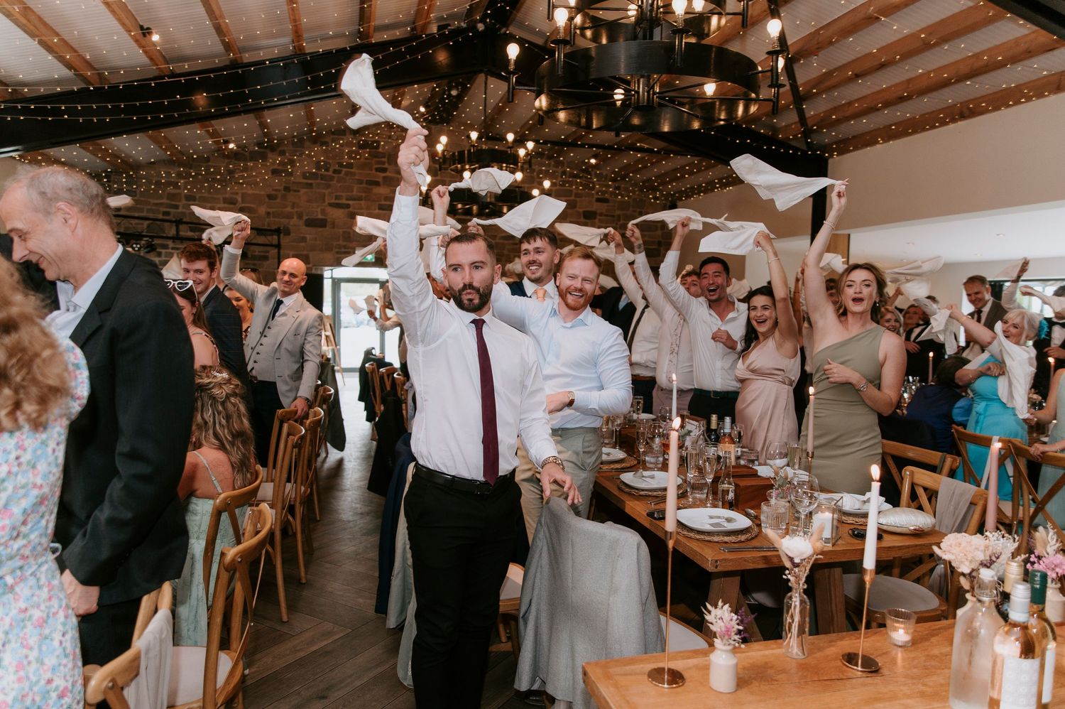 Wedding guests mingle and socialize at a rustic indoor reception venue with exposed wooden beams and warm lighting.