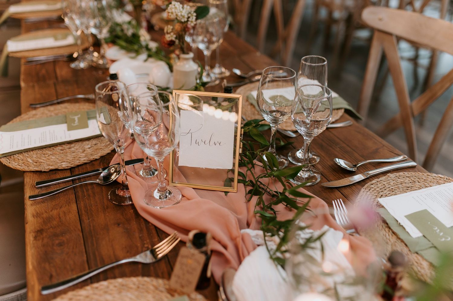 Rustic wedding reception table settings with blush pink floral centerpieces and wooden chairs in a natural light venue.