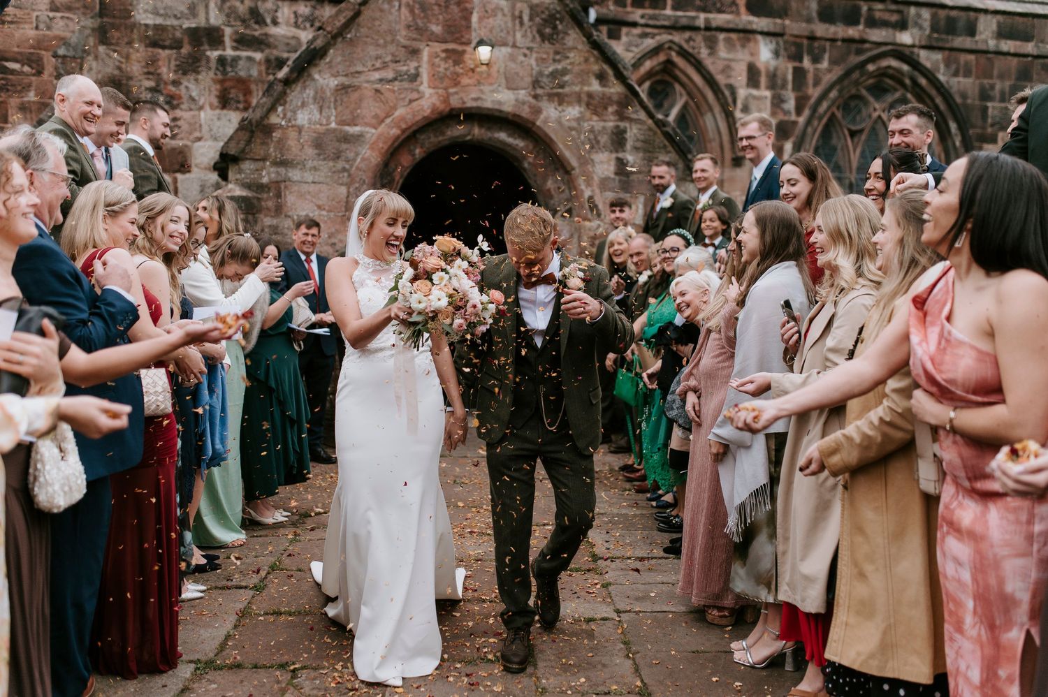 Wedding party celebrates in front of rustic stone wall as guests throw confetti during outdoor ceremony.