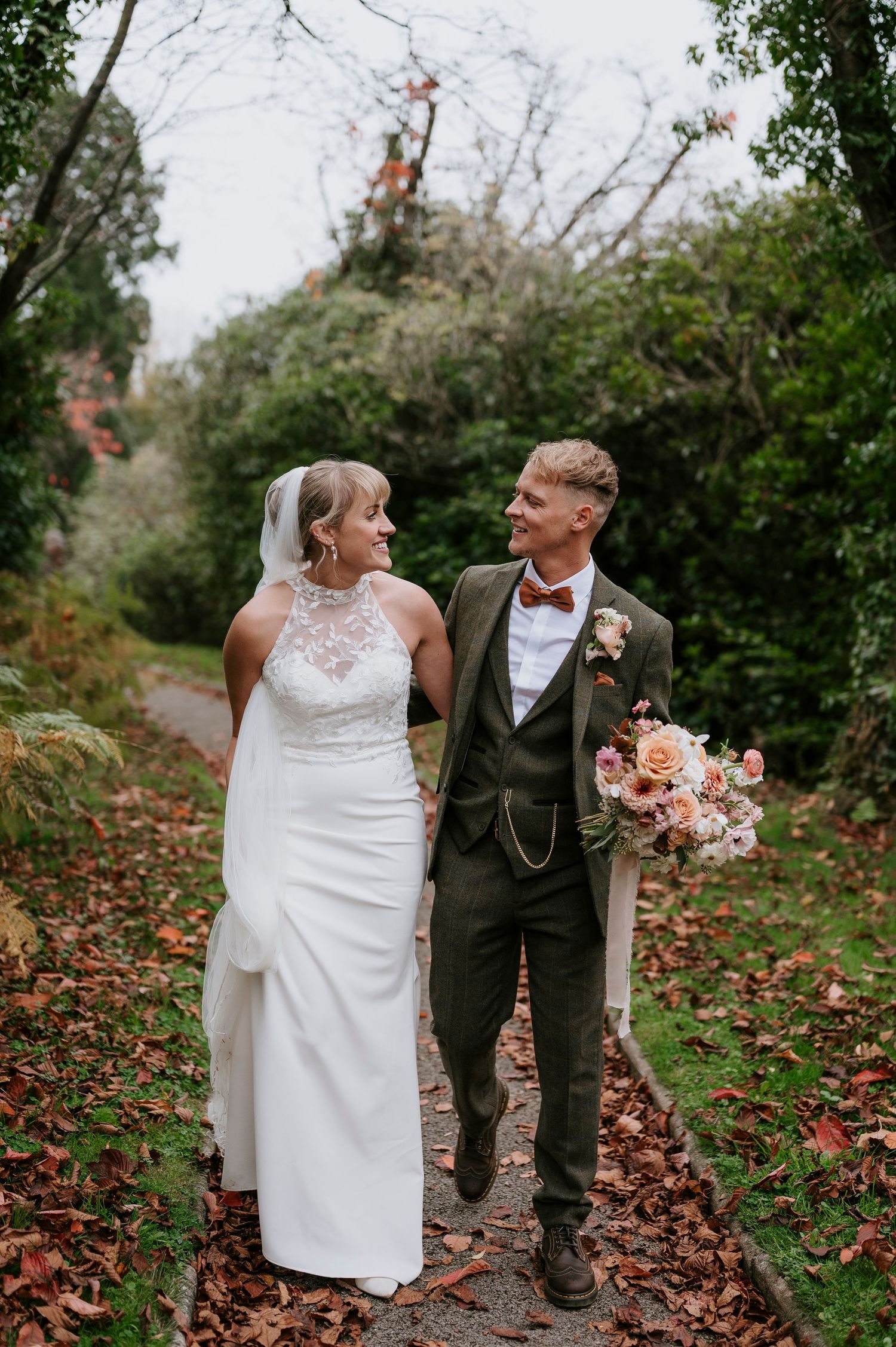 A couple in wedding attire walk and laugh together on a path covered in fallen autumn leaves.