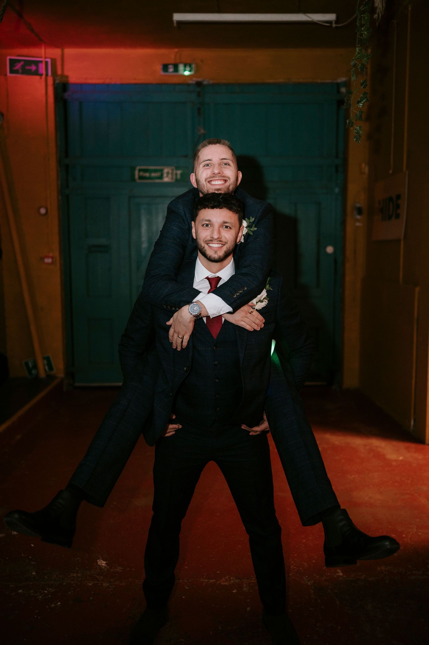 Two groomsmen in navy suits celebrating at wedding reception with piggyback pose near teal doors.