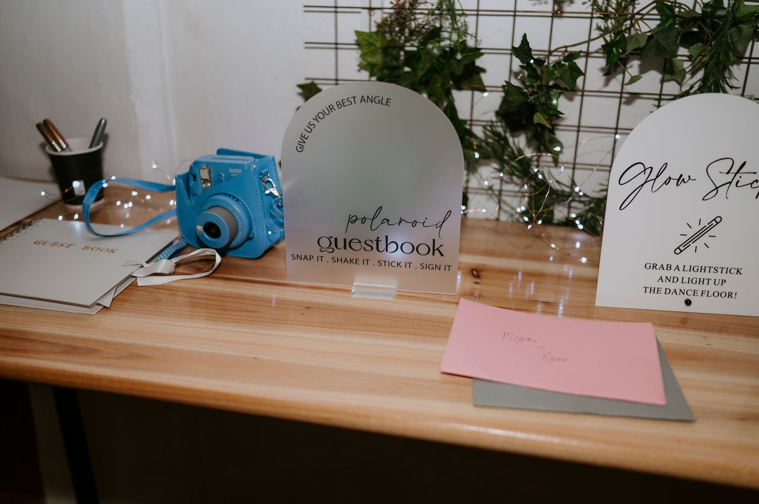 Blue Instax camera beside a polaroid guestbook sign and stationery on a wooden table at a wedding reception.