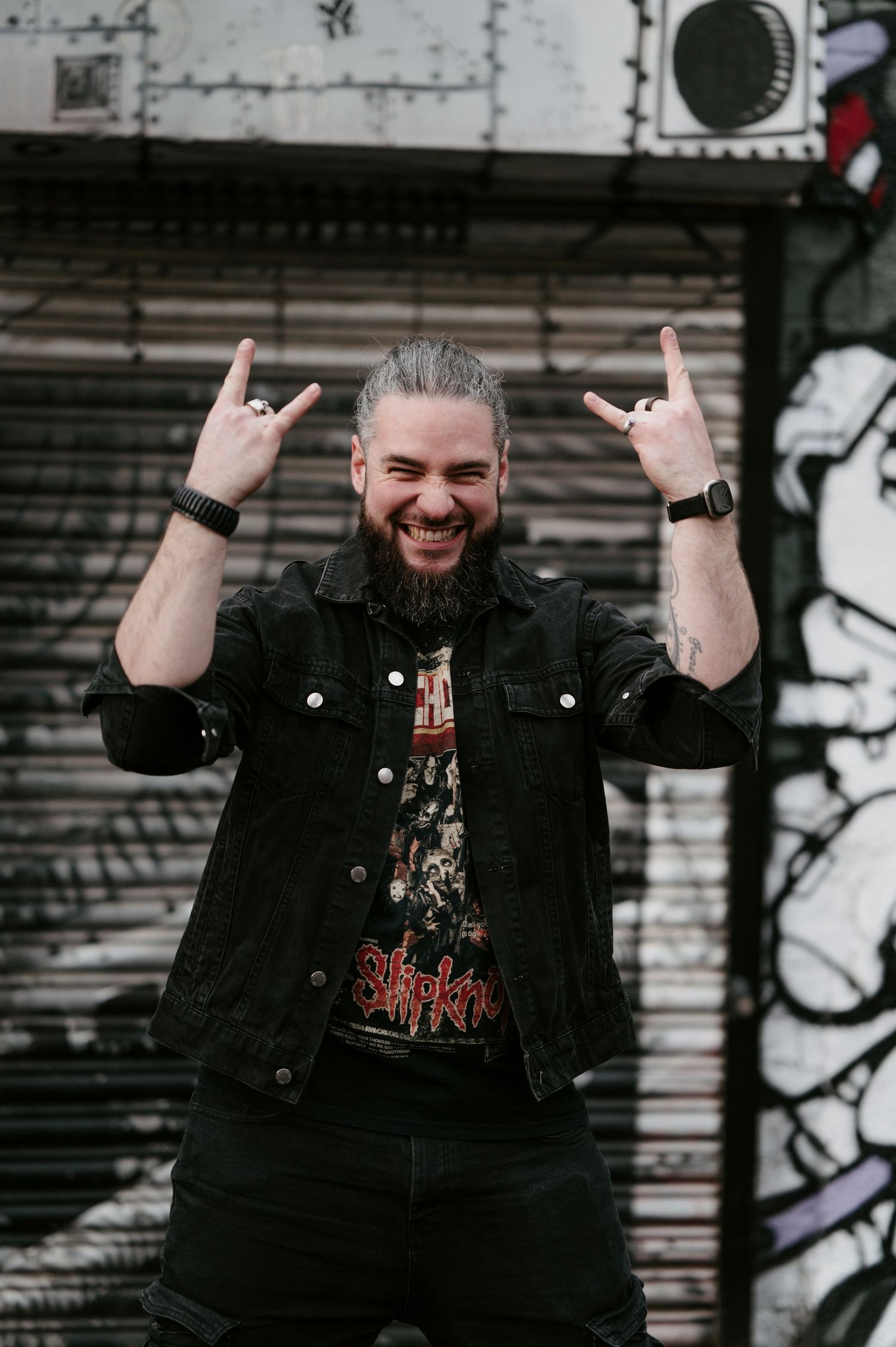 Man in black denim jacket shows enthusiasm with raised arms against graffiti wall background.