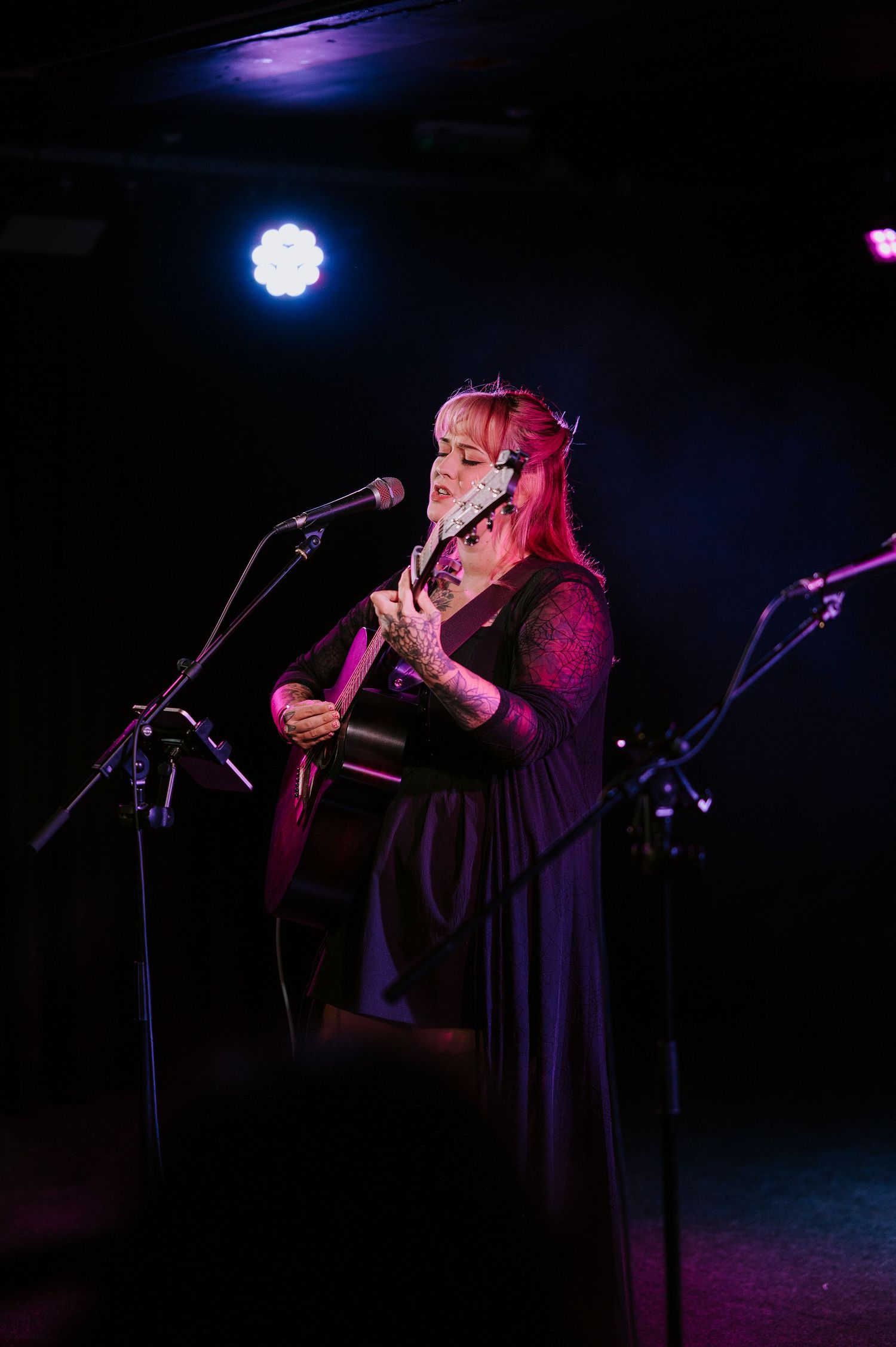 A performer in a dark dress sings passionately on stage under dramatic purple and blue lighting at a concert venue.