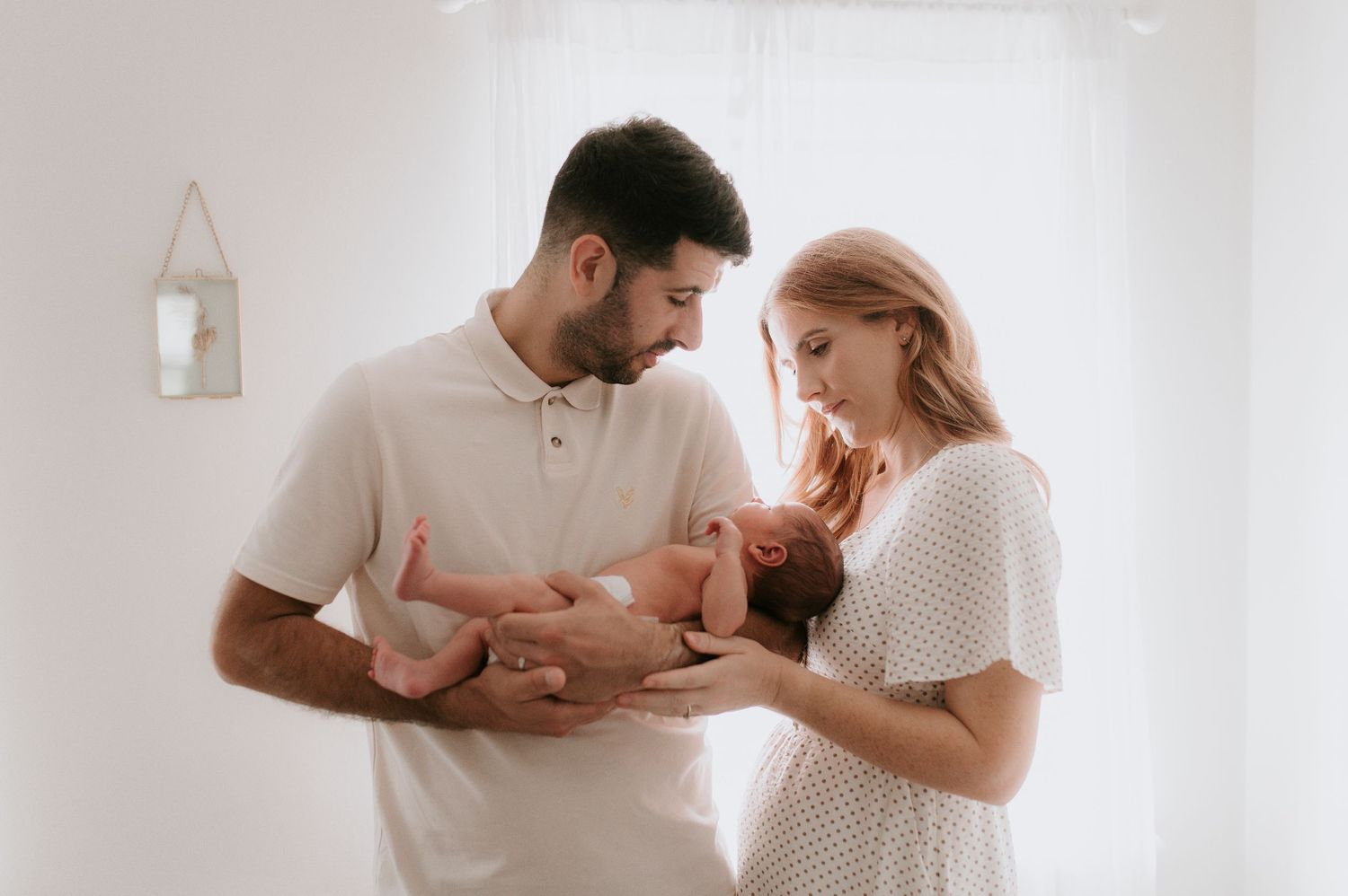 Parents in white outfits lovingly hold and smile at their newborn baby in a bright minimalist setting.