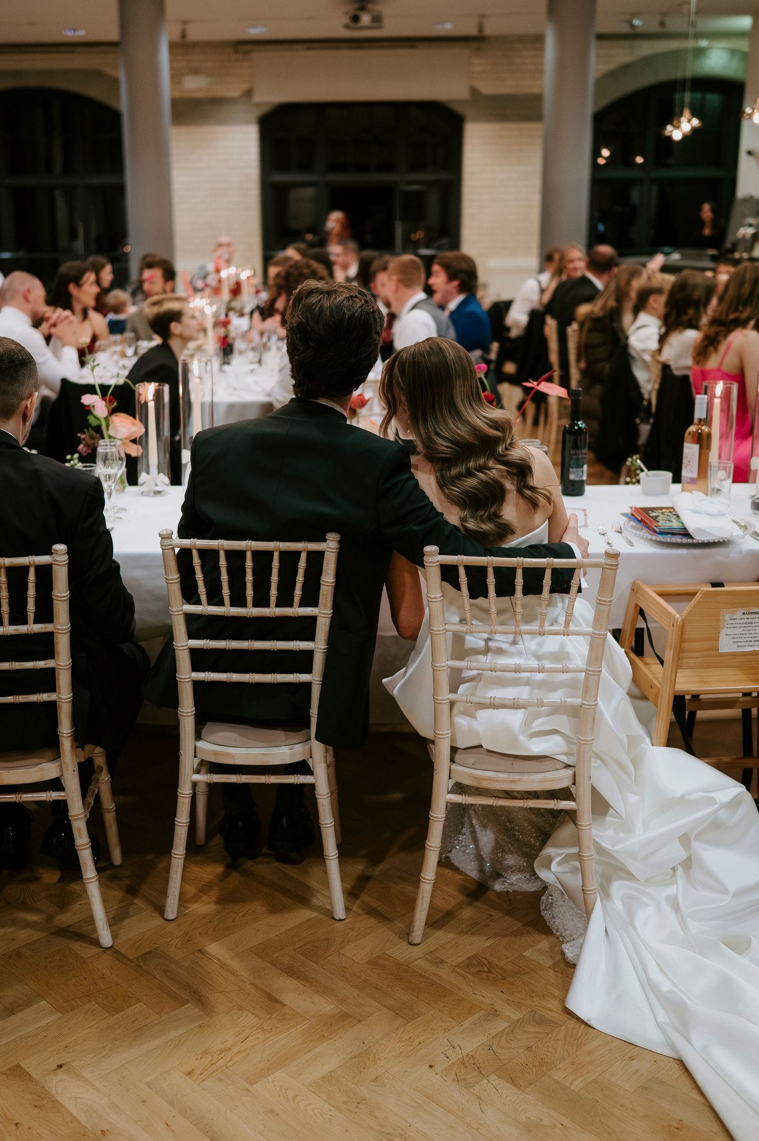 Wedding reception setup with white chiavari chairs and formal table settings on hardwood floors.