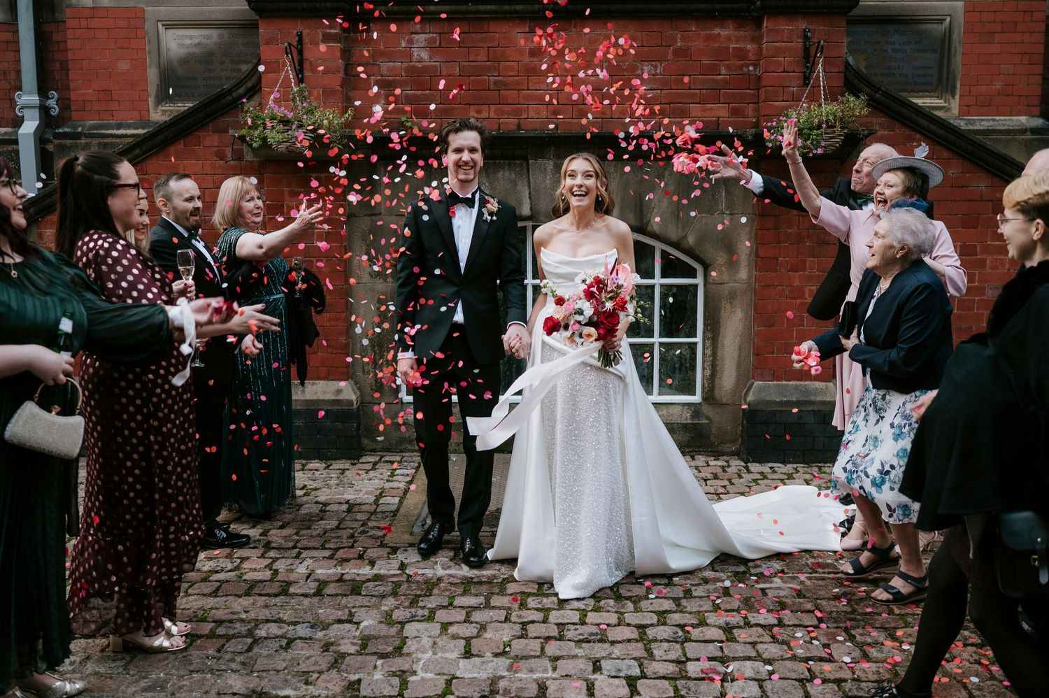 Wedding celebration on cobblestone street with guests throwing pink flower petals at couple outside brick building.