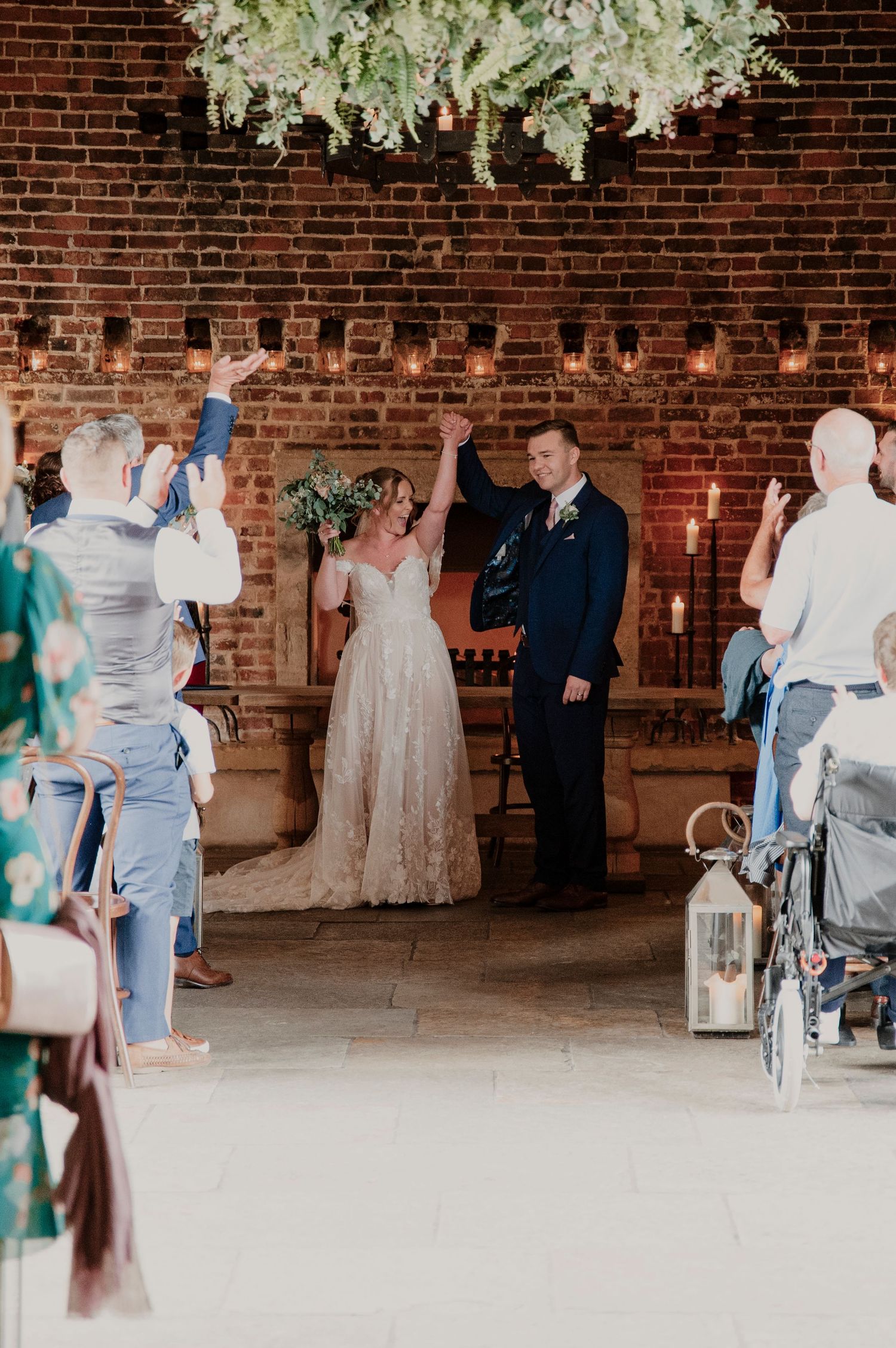 Wedding ceremony in a rustic brick venue with hanging white floral arrangements and candlelight decor.