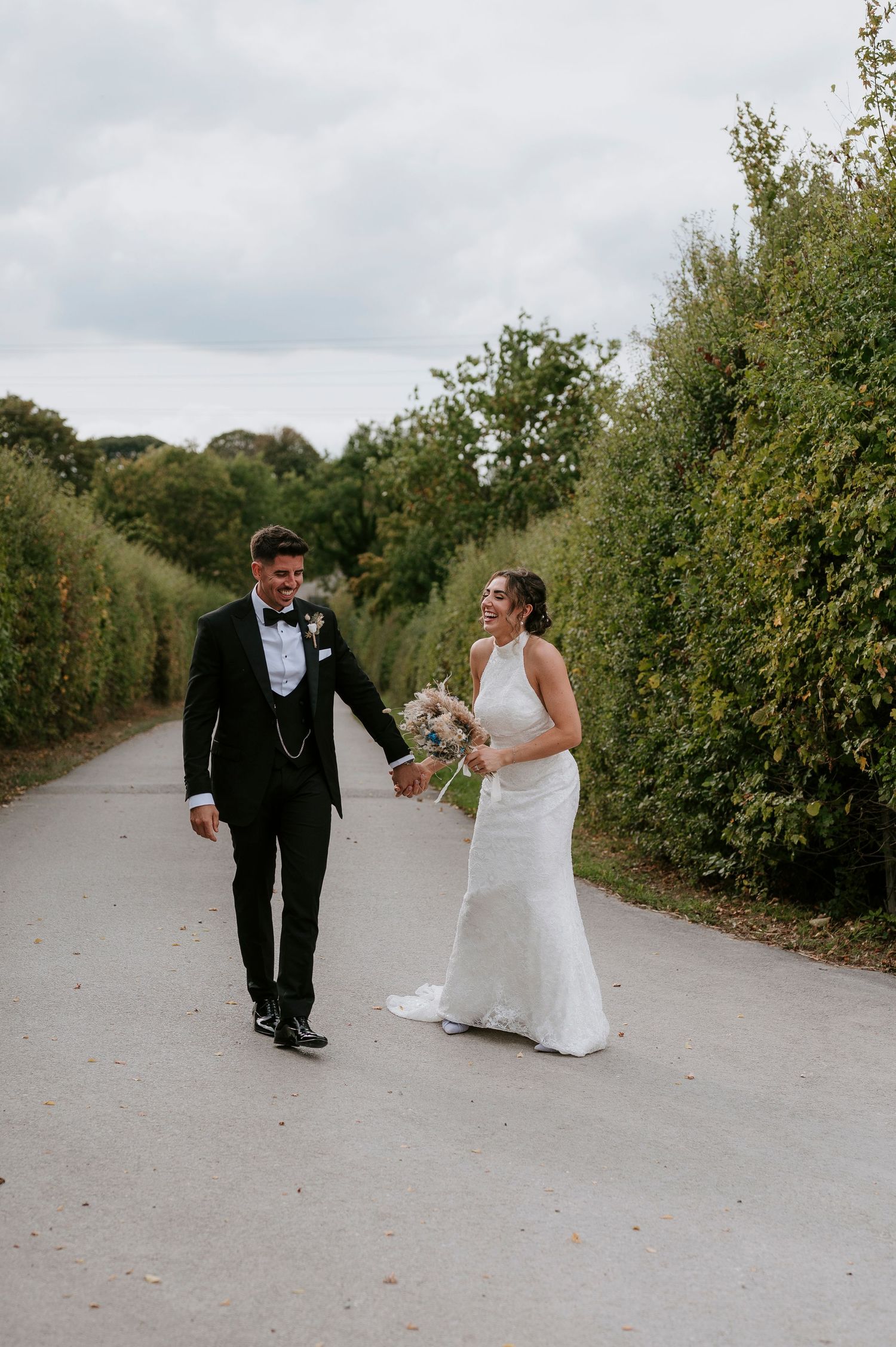 A couple in formal wedding attire playfully walks and dances together on a tree-lined path.