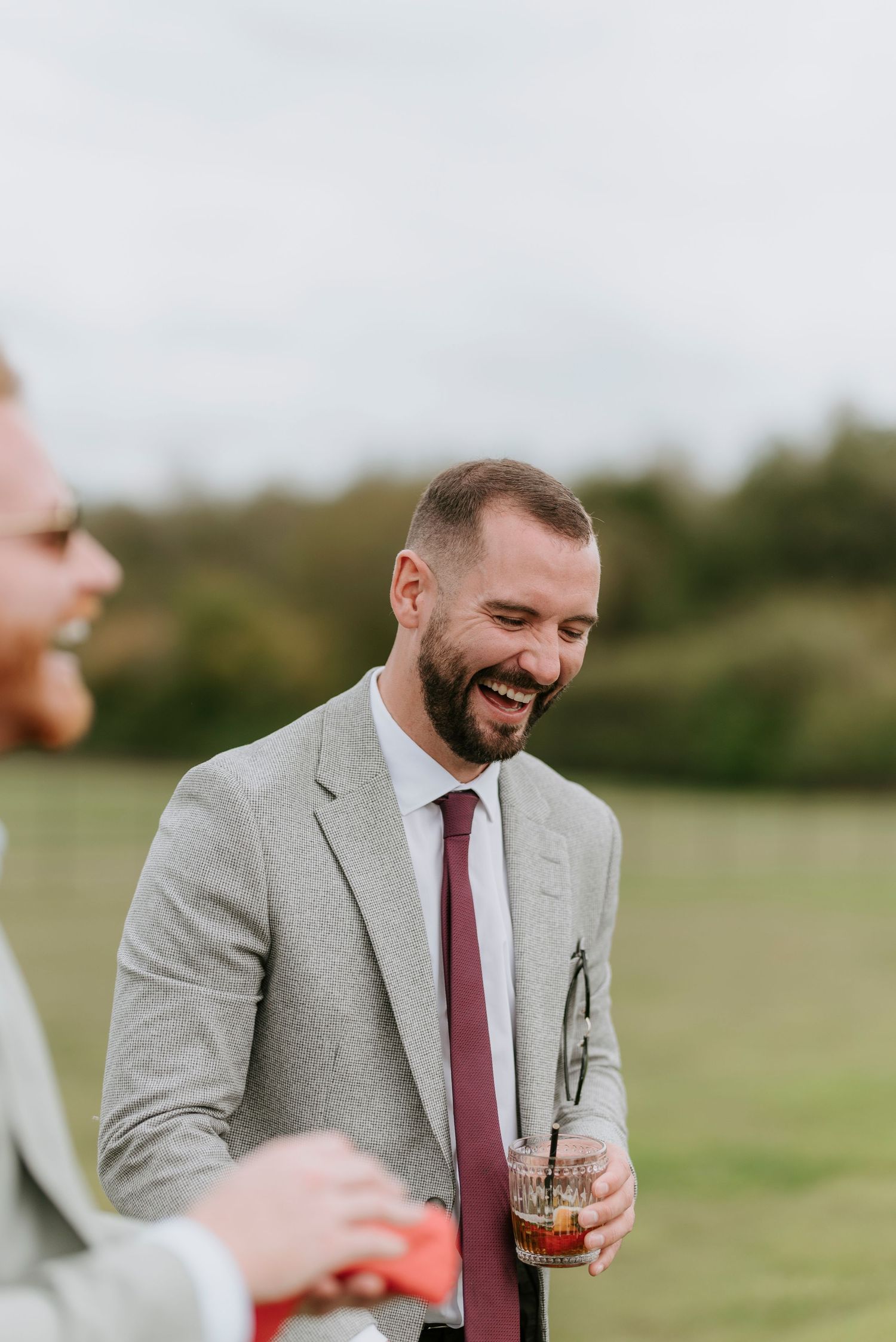 A person in a gray suit and burgundy tie laughs joyfully while holding a drink at an outdoor event.