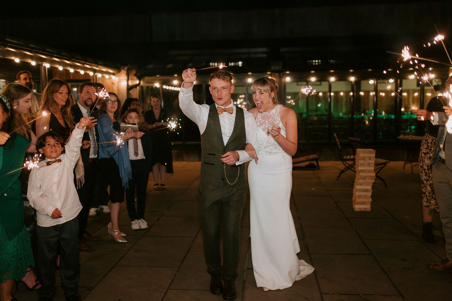 Wedding guests dance and celebrate on an outdoor patio decorated with string lights during an evening reception.