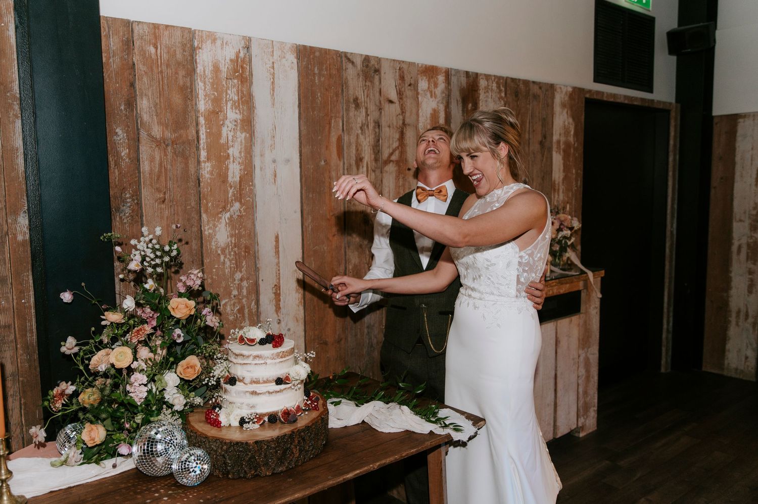 A wedding reception setup with rustic wooden walls, floral arrangements, and a white wedding cake on display.