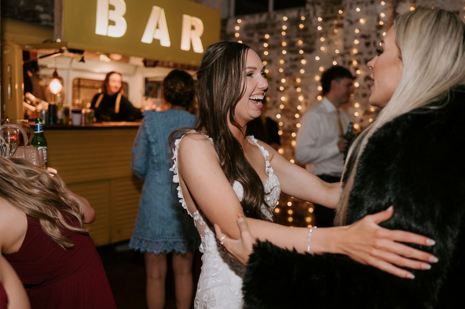 Bride in white lace dress laughing and dancing with guests near a decorated bar at a wedding reception.