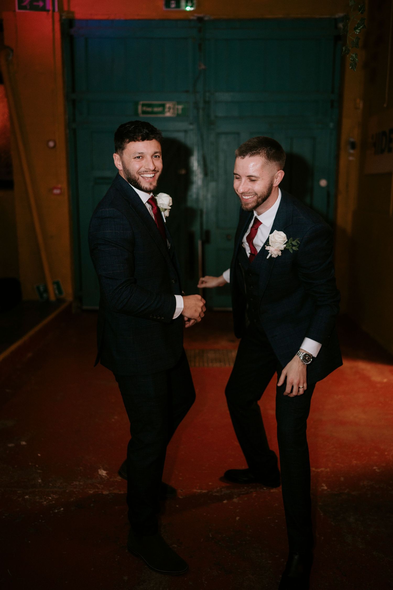 Two groomsmen in dark suits with white boutonnieres shaking hands and smiling at wedding reception.