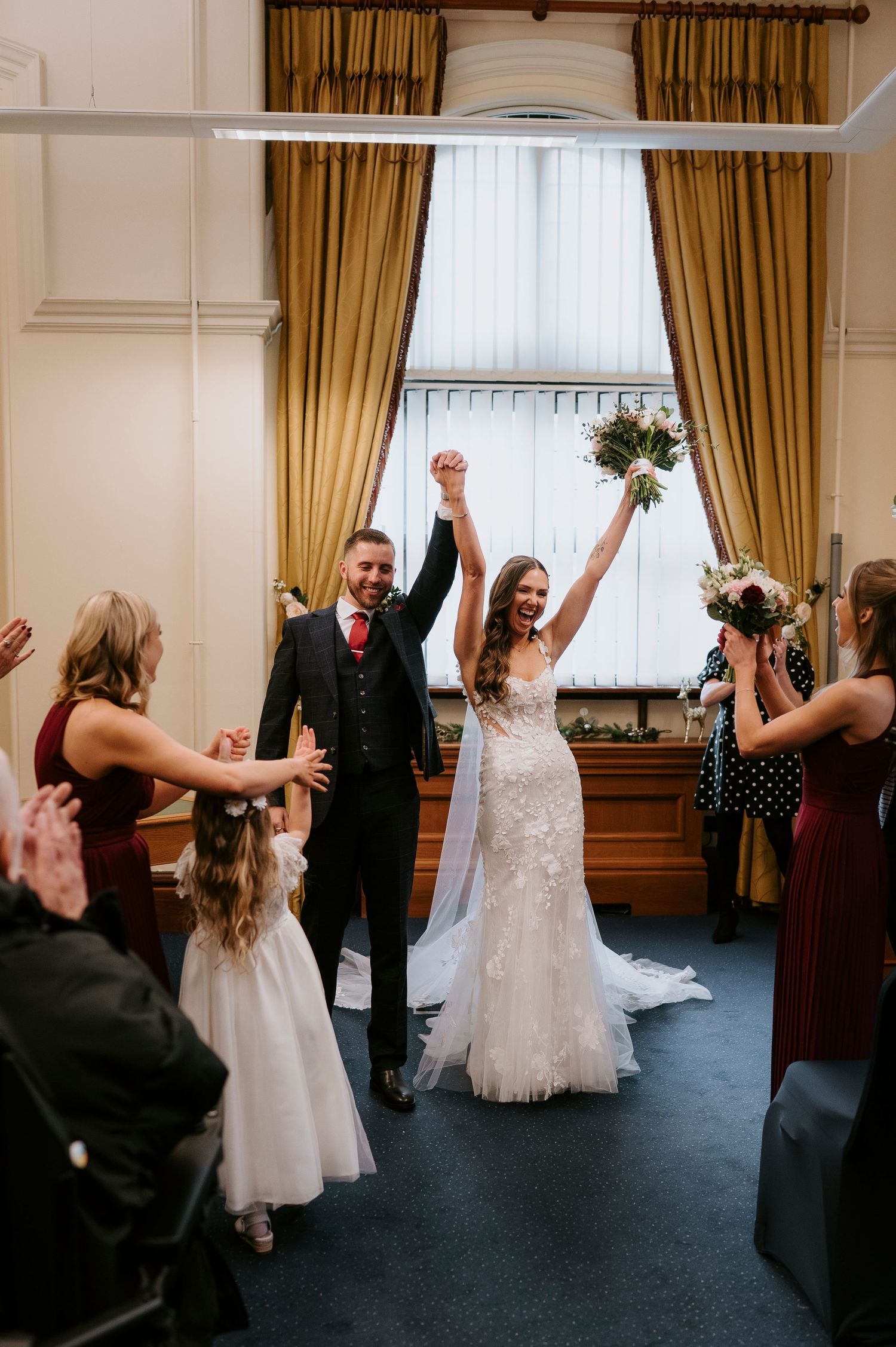 Joyful newlyweds raising hands after wedding ceremony in elegant room with gold curtains, bride in lace gown holding bouquet.