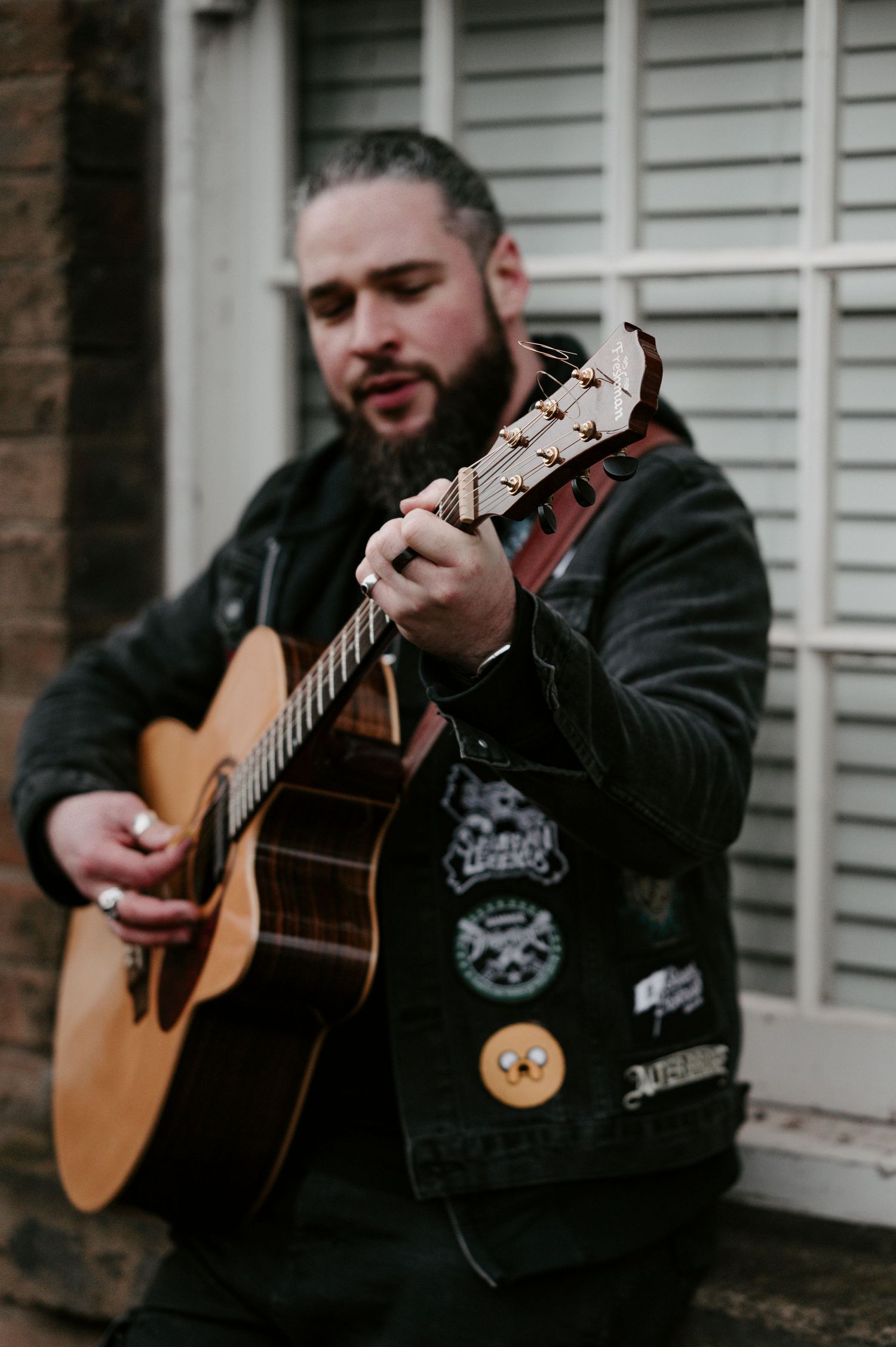 Musician in black leather jacket plays acoustic guitar against white siding wall outdoors.