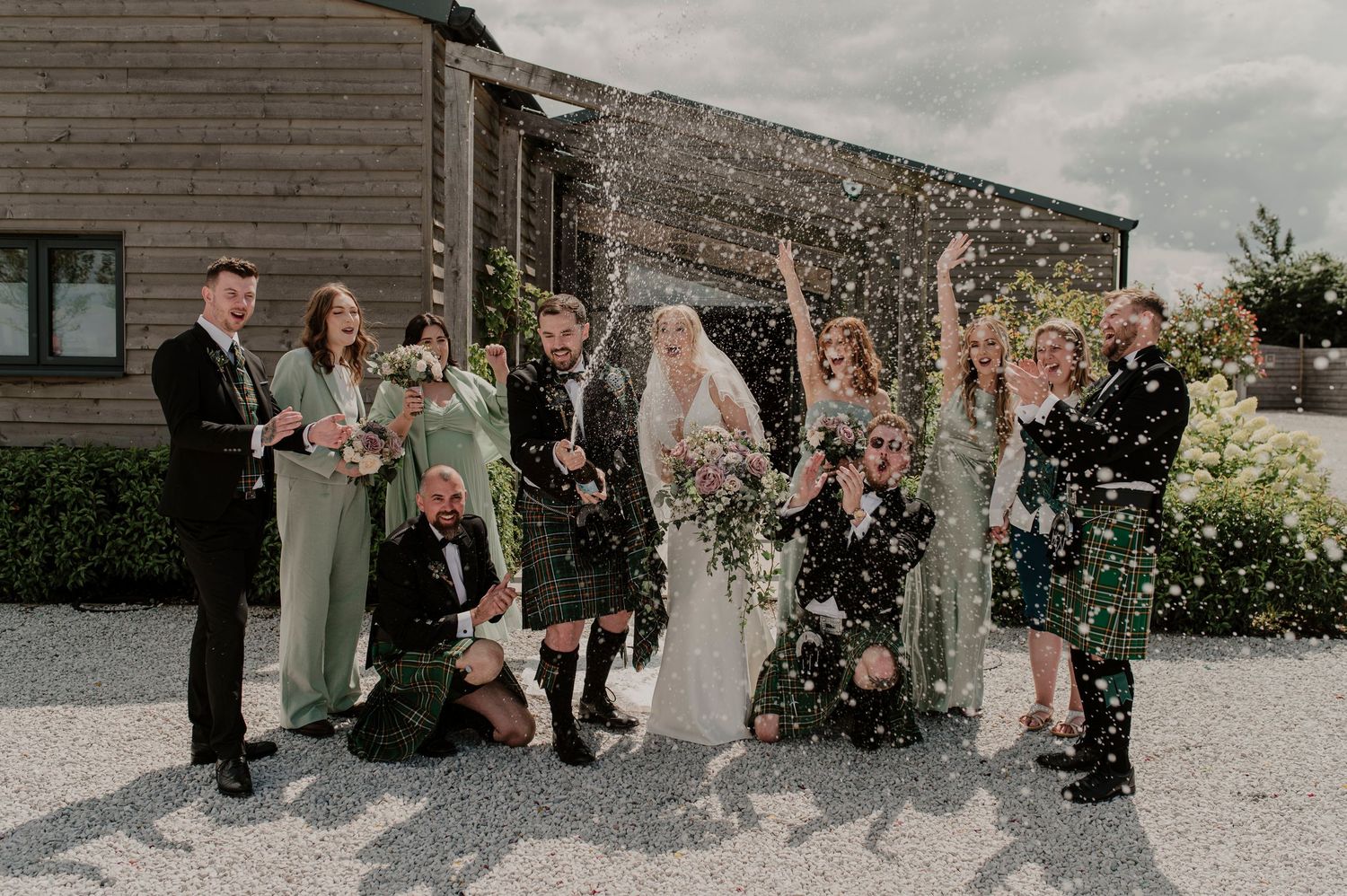 Wedding party celebrates with confetti toss outside stone building on overcast day.
