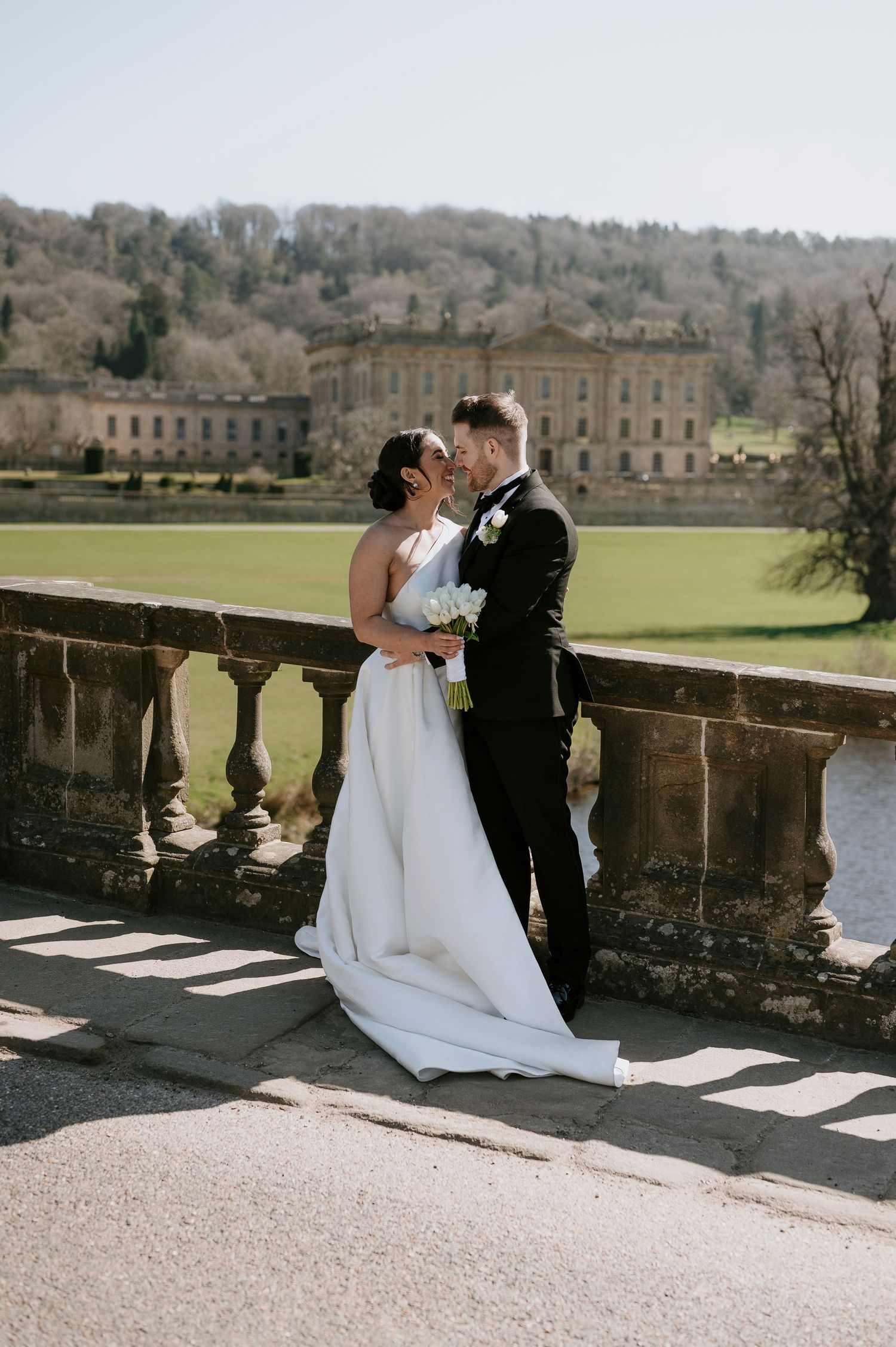 A romantic wedding couple embraces on a stone balcony overlooking the grounds of a grand historic estate.