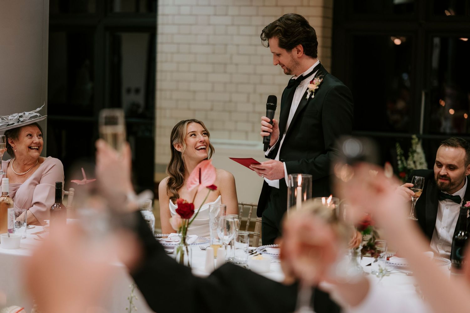 Wedding reception guests laugh and raise glasses during a heartfelt speech at an elegant indoor celebration.