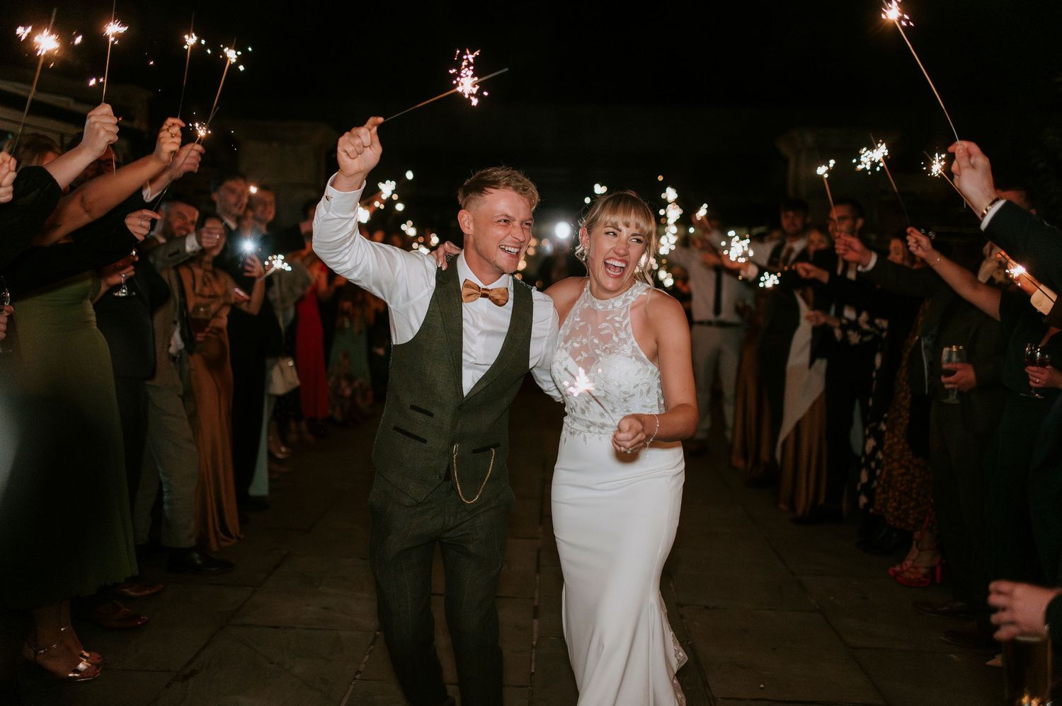 Newlywed couple joyfully exits their wedding reception through a sparkler tunnel lined with celebrating guests.