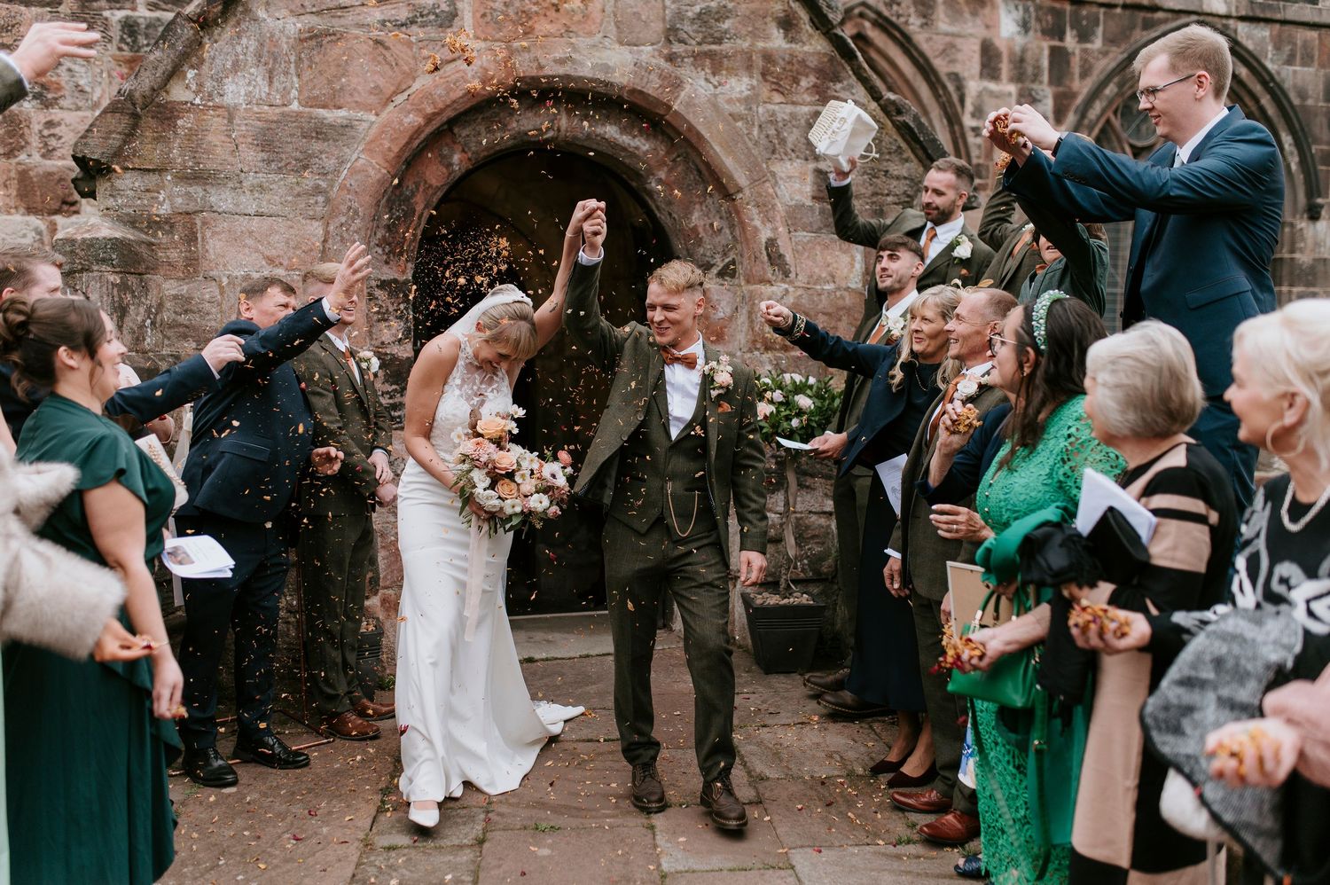 Wedding celebration with guests throwing confetti as couple exits stone archway at historic venue.
