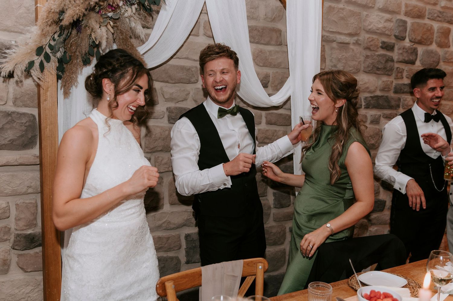 Wedding party members share joyful laughter during a candid reception moment at a rustic stone venue.