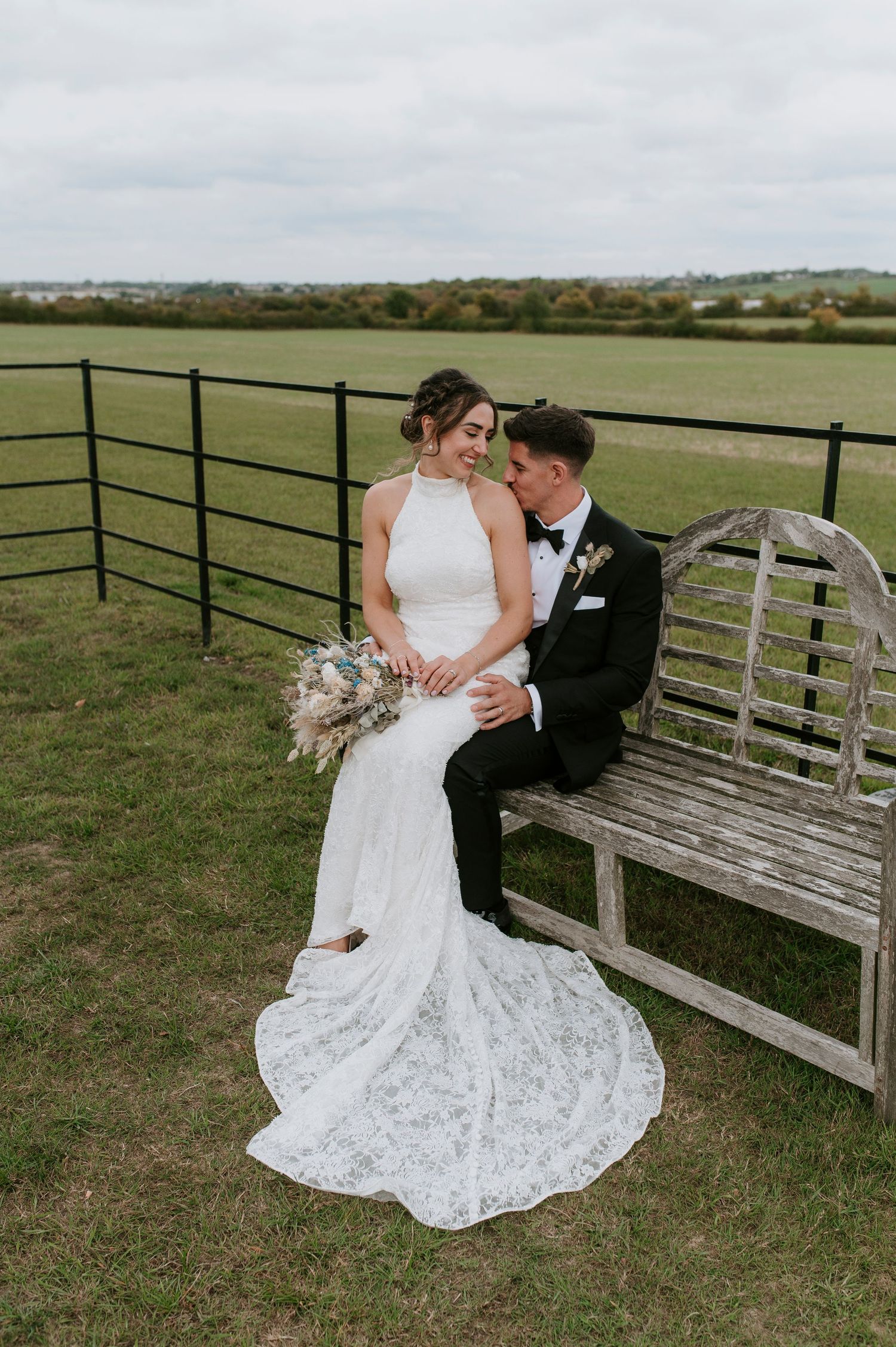 A couple in wedding attire sits intimately on a rustic wooden bench overlooking green countryside fields.